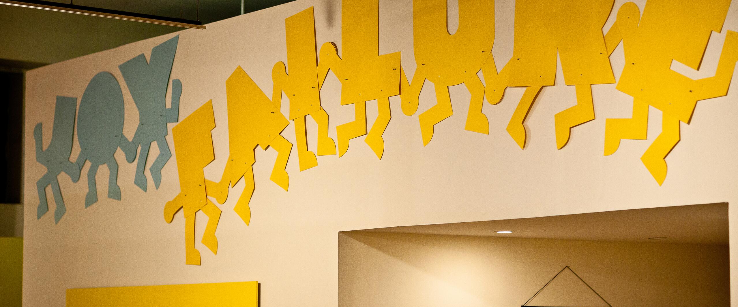 Colorful wall art with stylized figures spelling "JOY FAILURE" in blue and yellow letters. Below, a yellow rectangle displays fragmented text. The setting is an indoor gallery with muted lighting.