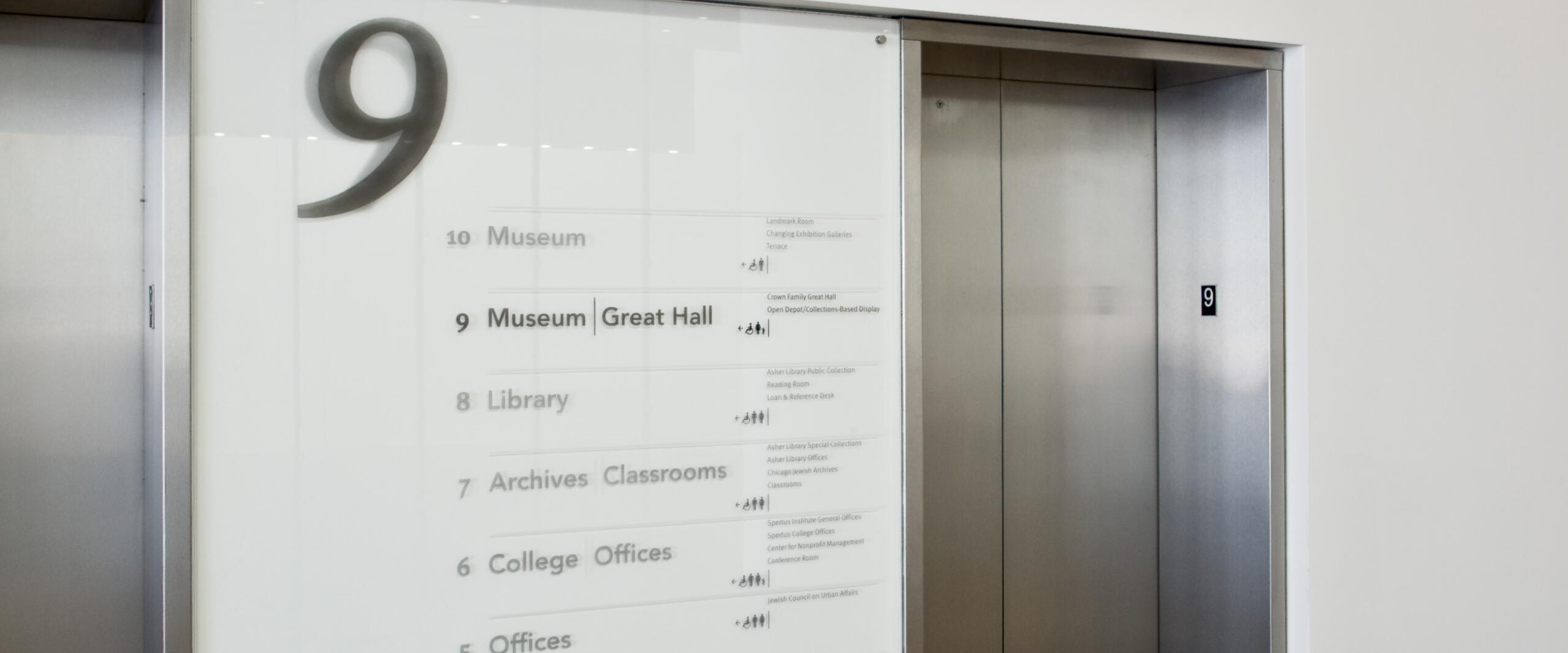 Elevator lobby with a floor directory sign showing different levels, such as Museum, Library, and Children's Center. Two metal elevator doors with the number nine above them. Wall text reads "Bernard & Rochelle Zell Holocaust Memorial."