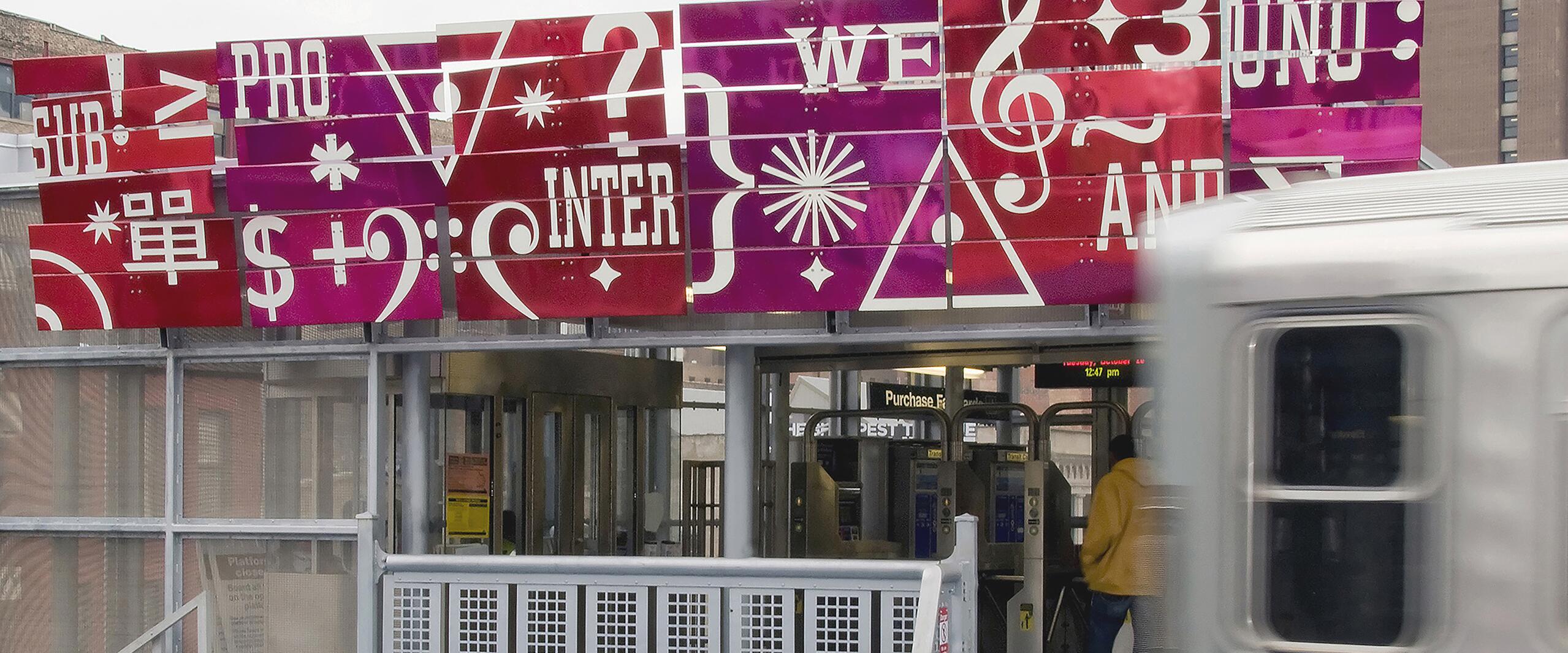 Colorful mural with various symbols and words above a train station entrance, a train passing by in the foreground. A person in a yellow jacket is entering through the turnstile. Tall buildings in the background.