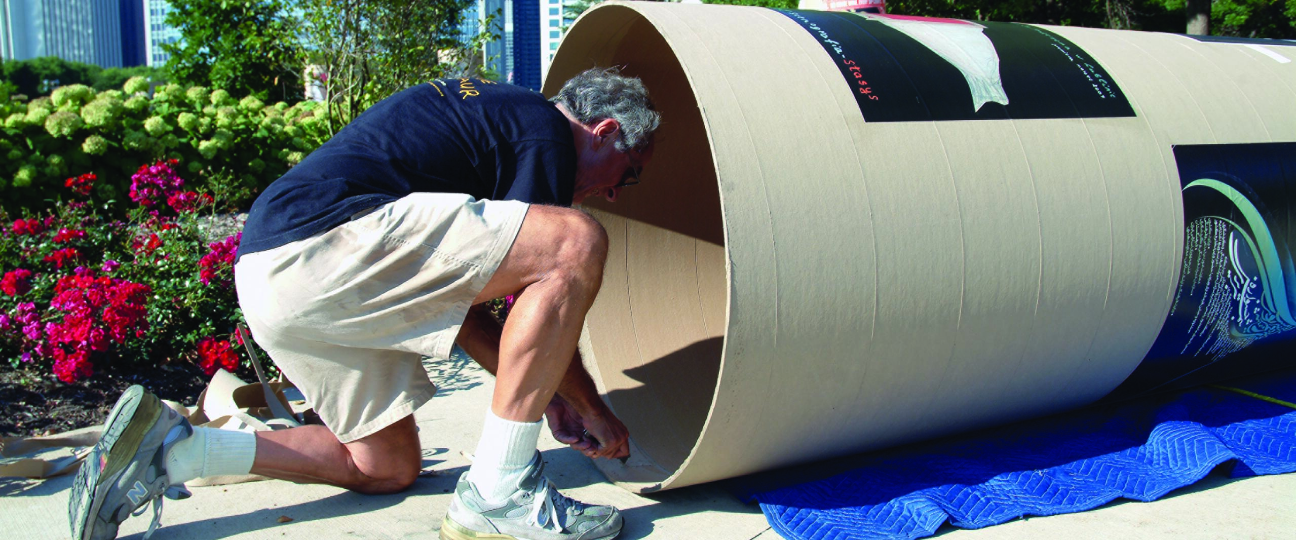 Man wearing shorts and sneakers kneels beside a large cylindrical cardboard structure on a sidewalk in an urban park. He is working on the structure, surrounded by blooming flowers and city skyscrapers in the background.