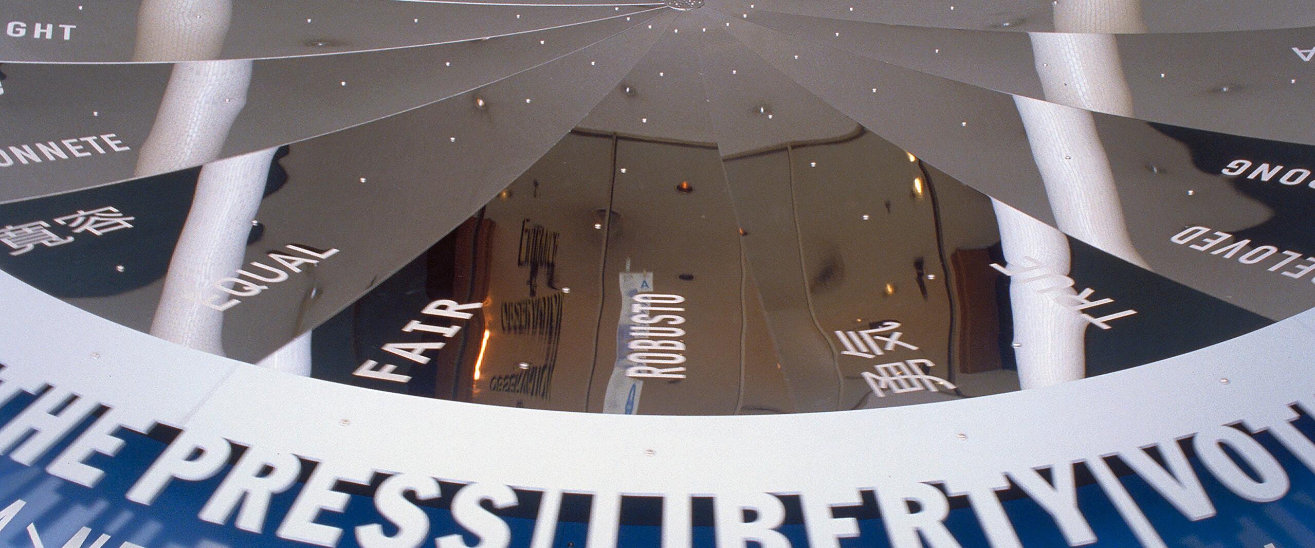 Circular ceiling with reflective metallic panels displaying words in various languages, including "LIBERTY" and "NEVER FORGET." Lights and reflections create a dynamic visual effect.