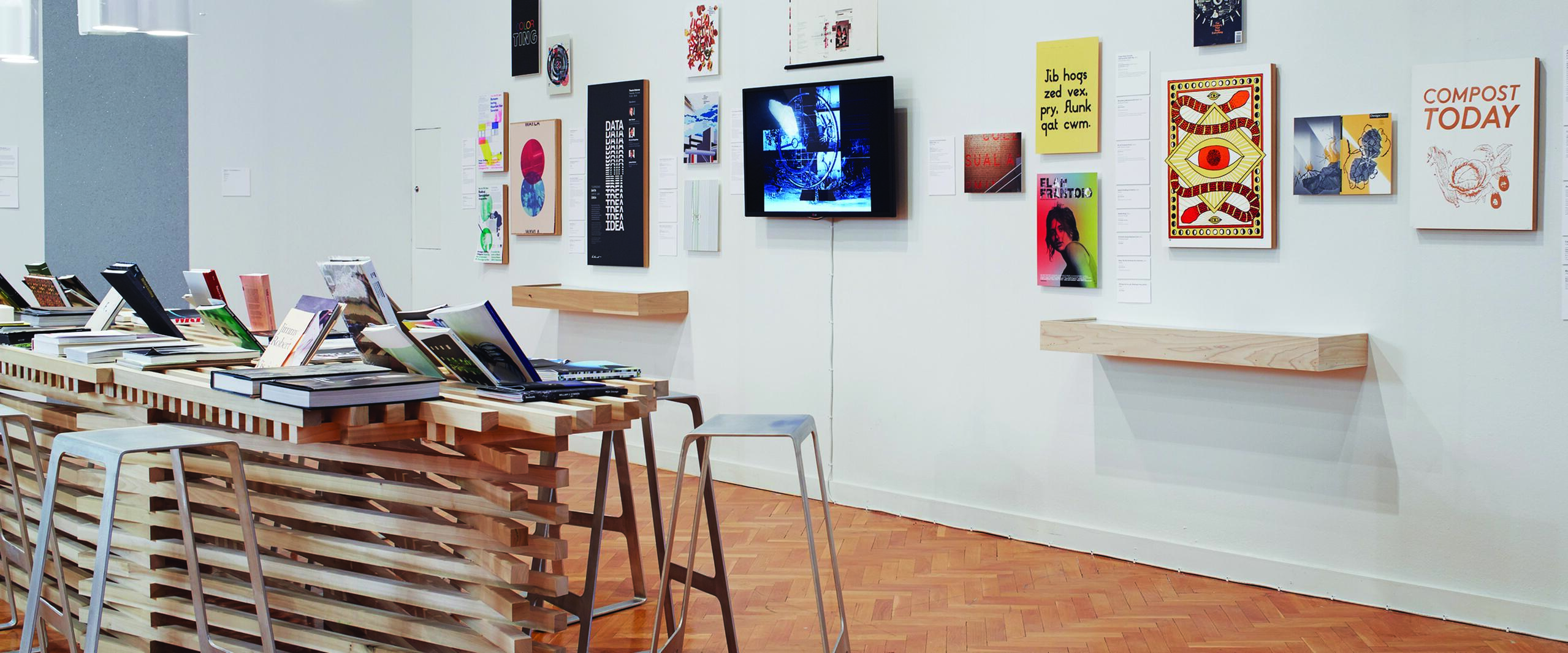 Modern gallery with books on a wooden table, stools, and eclectic artwork on white walls. A screen exhibits digital content. Bright, minimalist lighting hangs above.