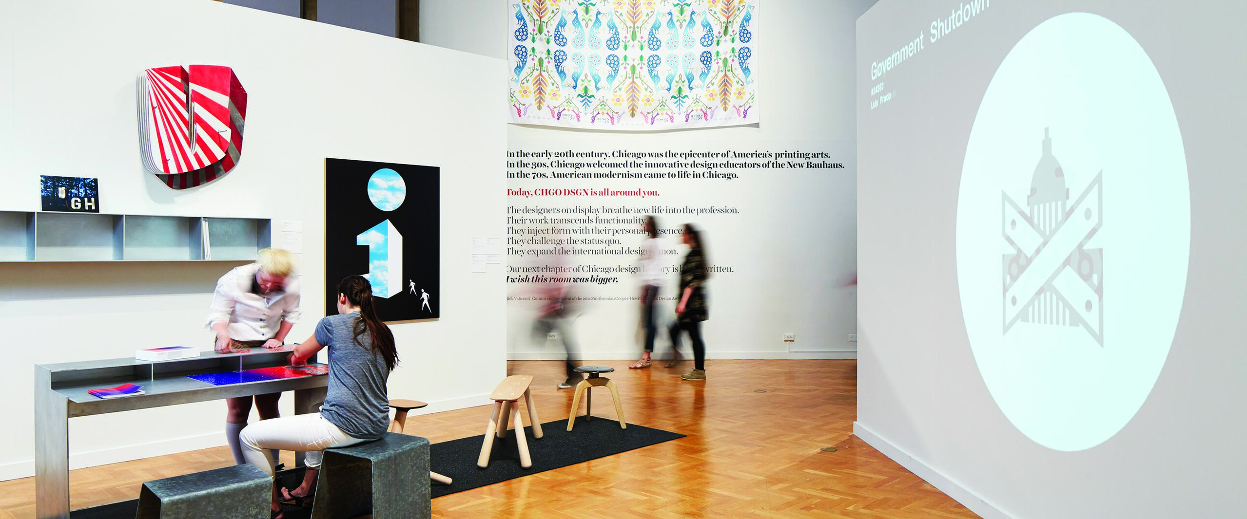 Exhibit scene with two people interacting at a modern metal table. Artwork and graphic elements adorn the walls, while text and a projected image are visible. Wooden parquet flooring and stools provide a contemporary setting.