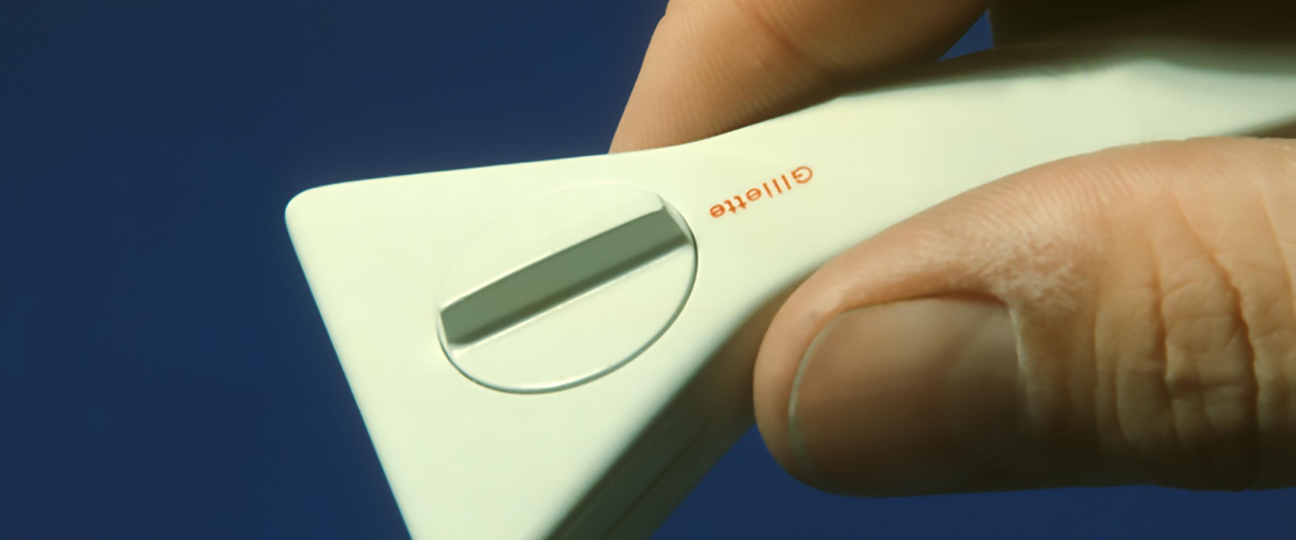 Hand holding a white Gillette razor with a triangular shape and circular blade cover, set against a blue background.