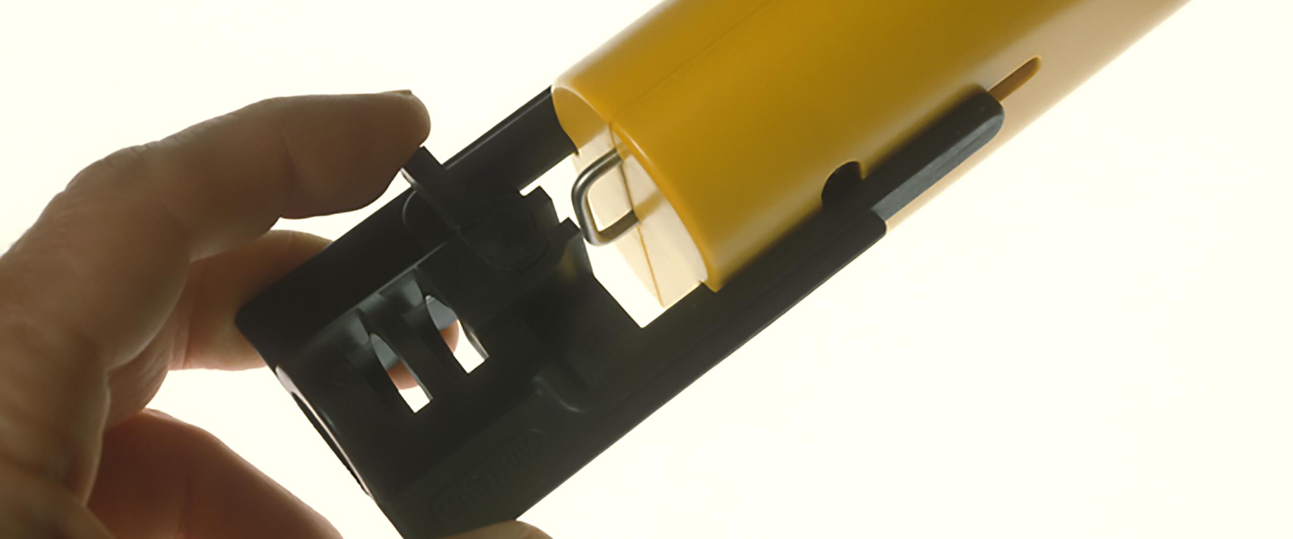 A hand holding a black and yellow plastic device, revealing its internal mechanism with a visible wire loop, set against a light background.