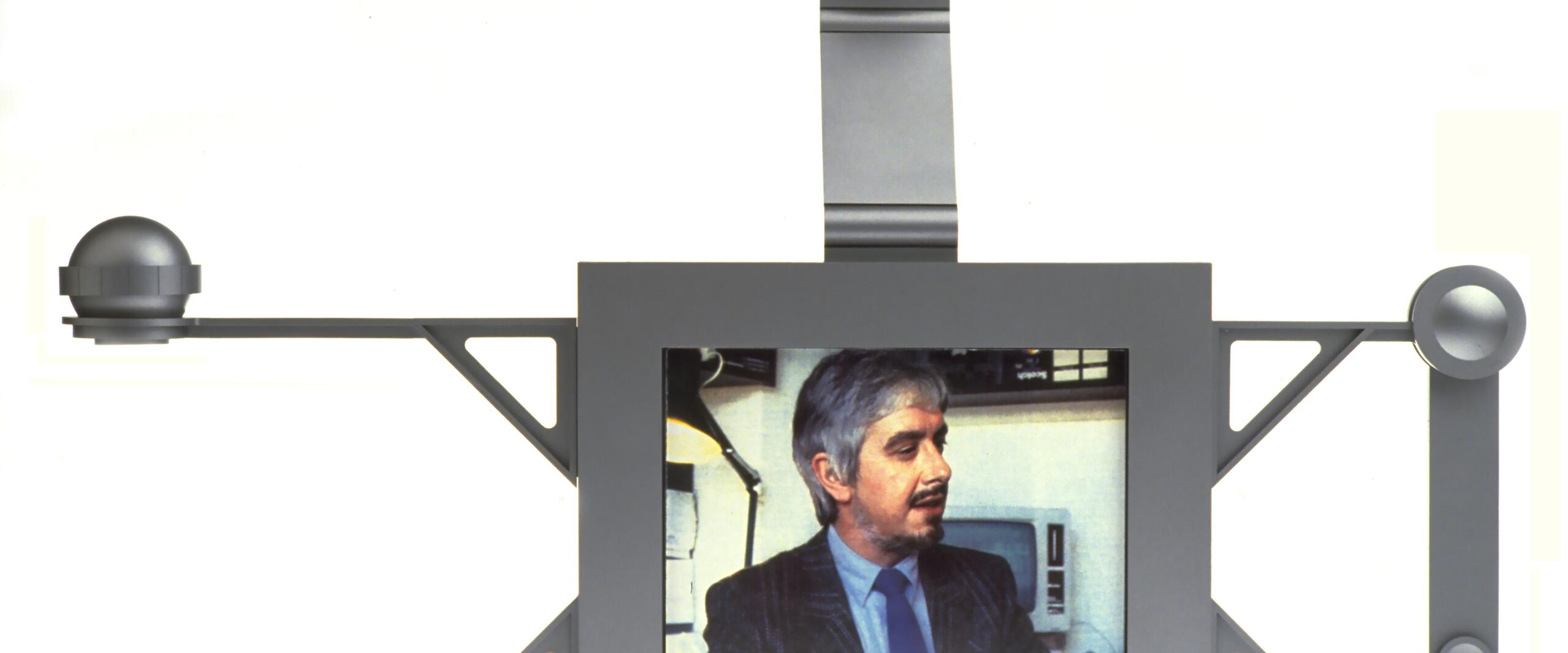 Futuristic video conferencing device with a central screen showing a man in a suit, flanked by angular supports and round sensors on both sides, topped by a camera.