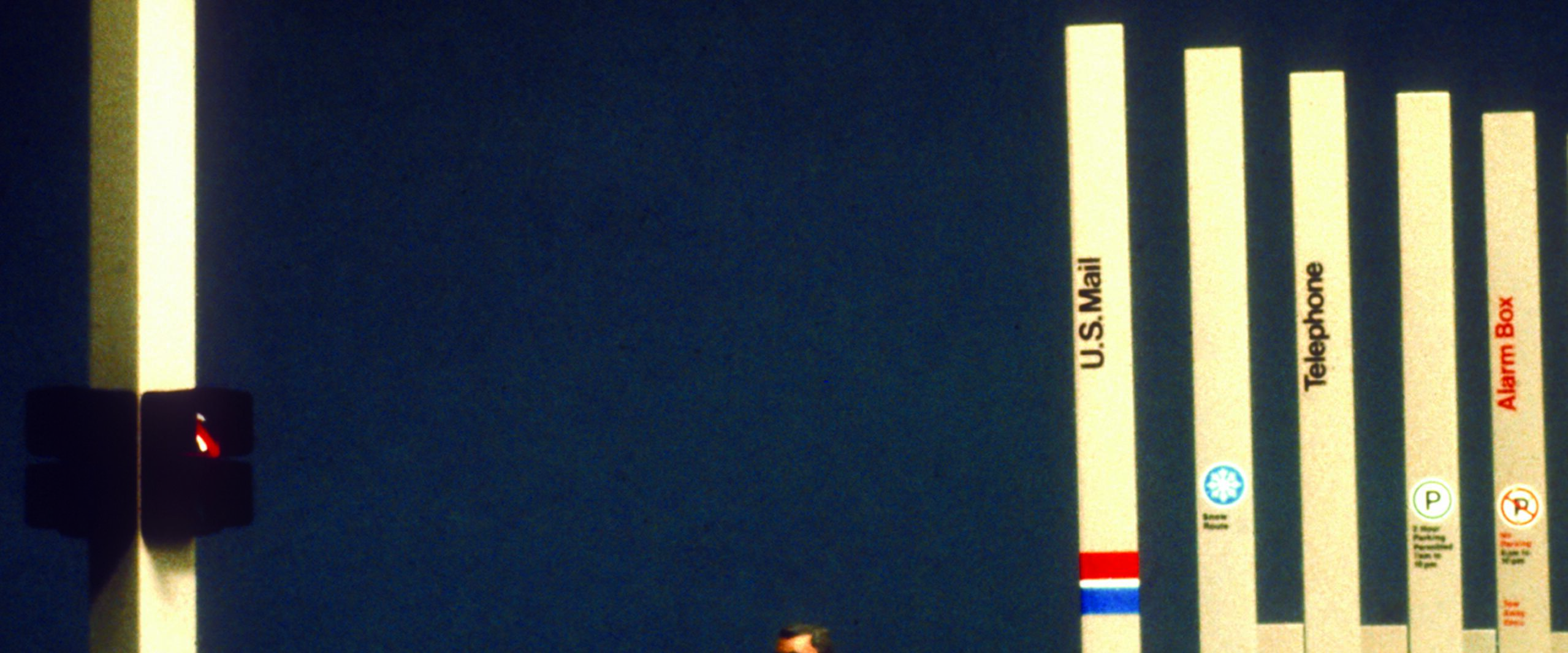A man in a suit walks past a series of tall, vertical signs including U.S. Mail, Telephone, and Alarm Box. A traffic light shows red. The street sign reads "Lexington Av" with directional arrows. The background is dark, enhancing the illuminated signs.