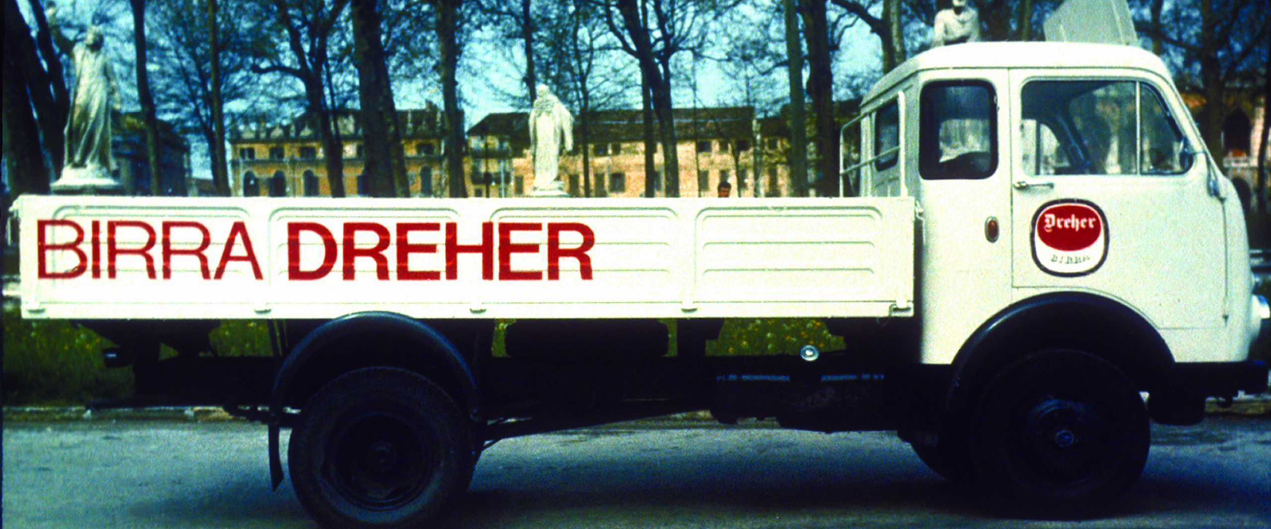 White delivery truck with "Birra Dreher" in red on the side, parked in a tree-lined area. Plaque with a similar design on the driver's door. Background features historic buildings partially visible through the trees.