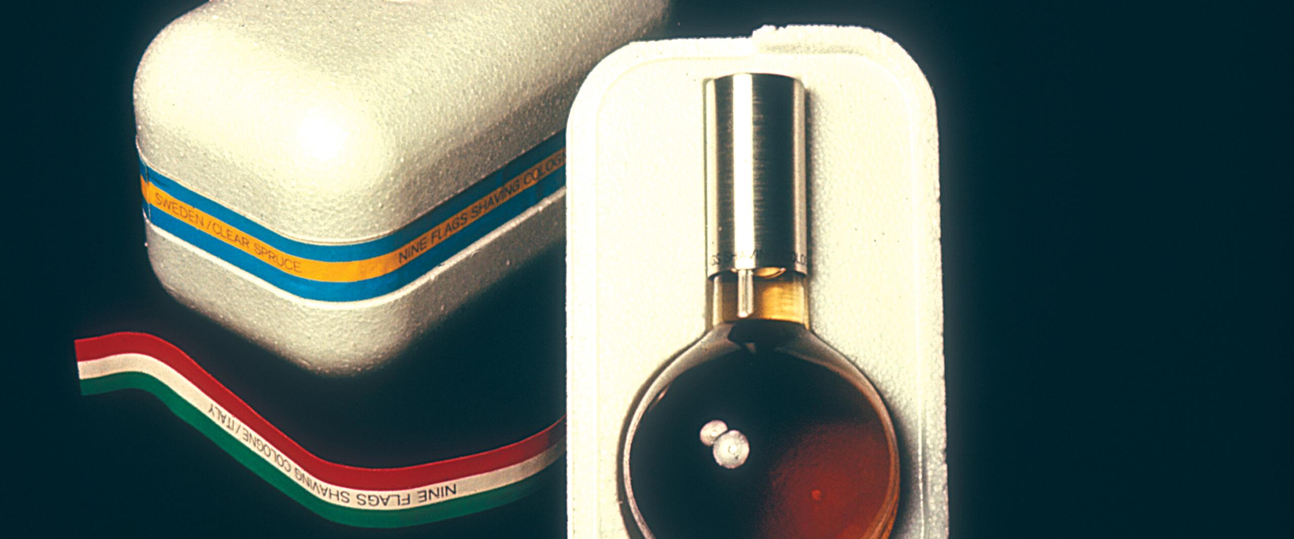 Cologne bottle with silver cap in a white foam container, featuring a striped box with text and tricolor ribbon.