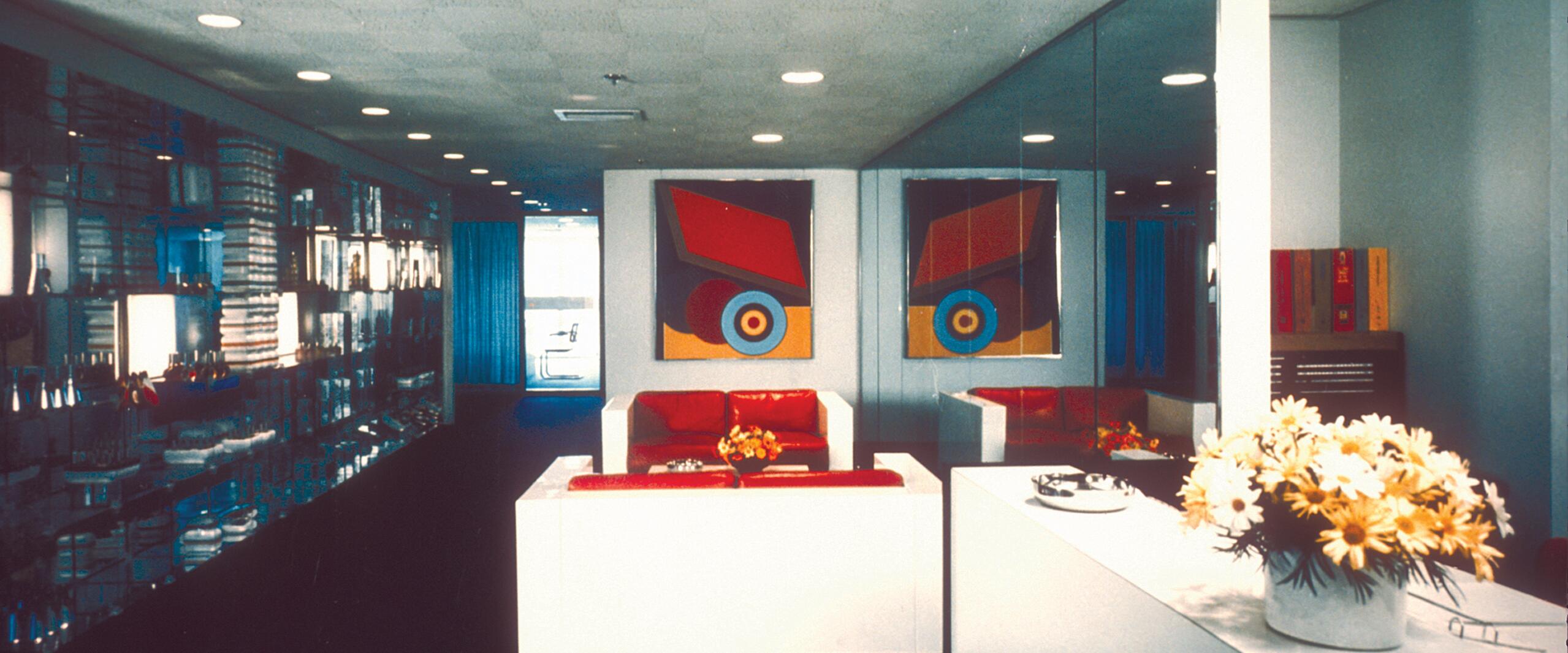 Modern lobby with mirrored walls, red seating, and abstract art. White counter with a vase of yellow flowers. Shelves on the left display items, and natural light filters through a window in the back.