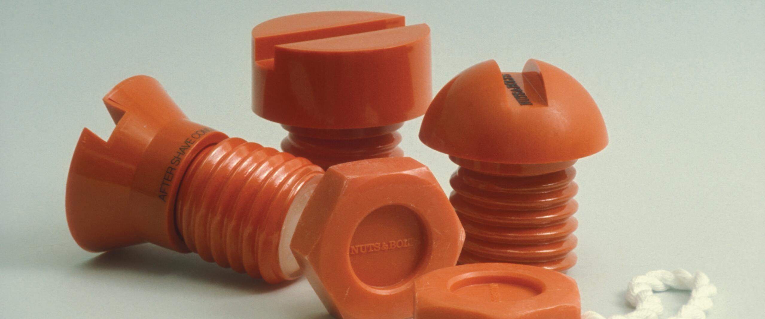 Orange plastic objects resembling oversized screws and bolts arranged together on a light background. A white rope is attached to one piece.