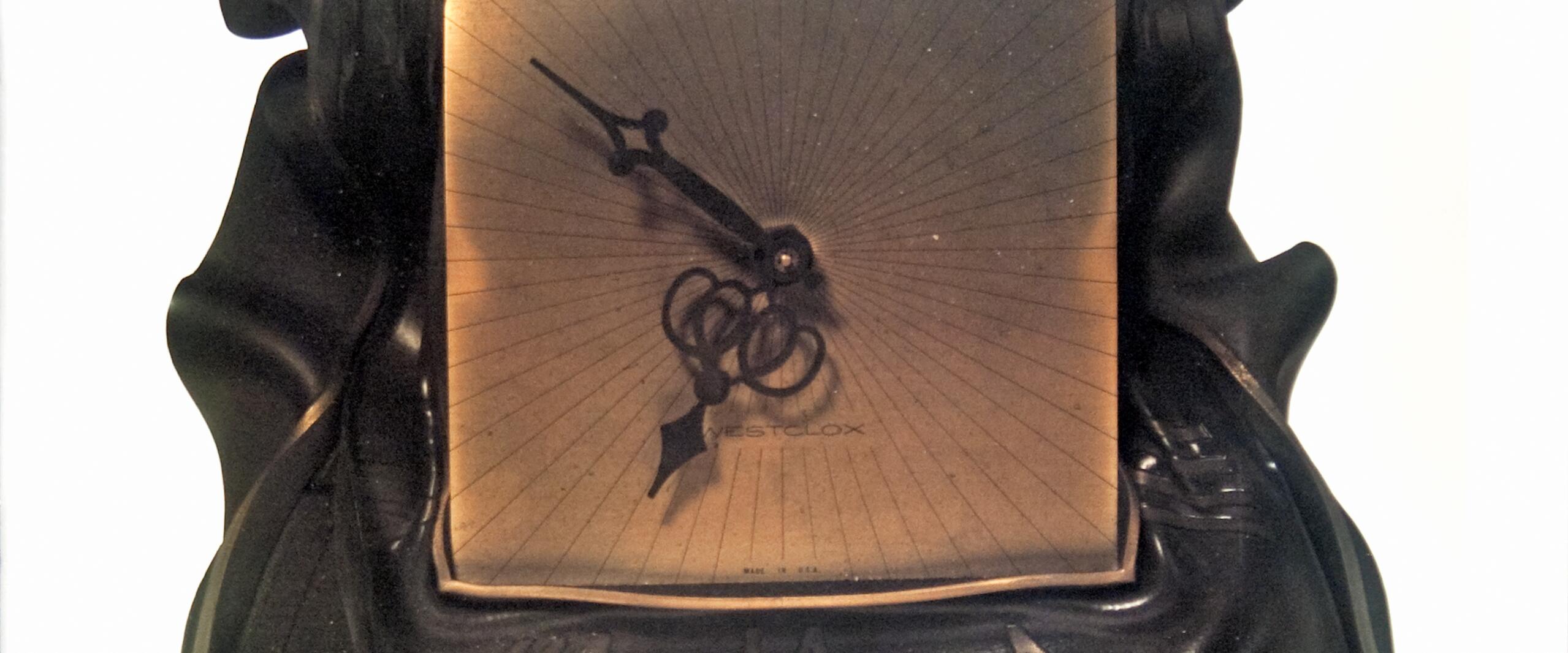 A square bronze sundial with ornate hands on a draped black fabric background.