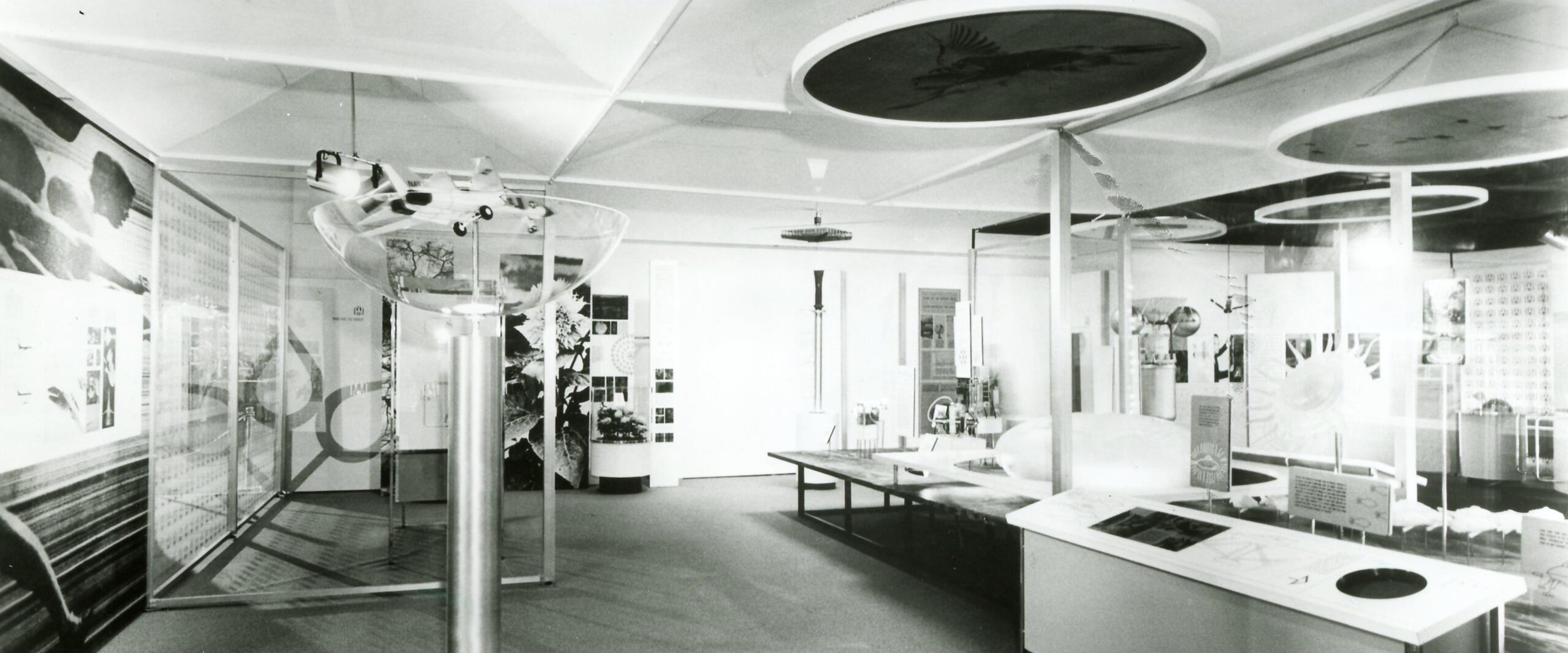 Black and white photo of a modern exhibition room with displays on aerodynamics. Circular models hang from the ceiling, and a variety of educational panels and diagrams are on tables and walls.