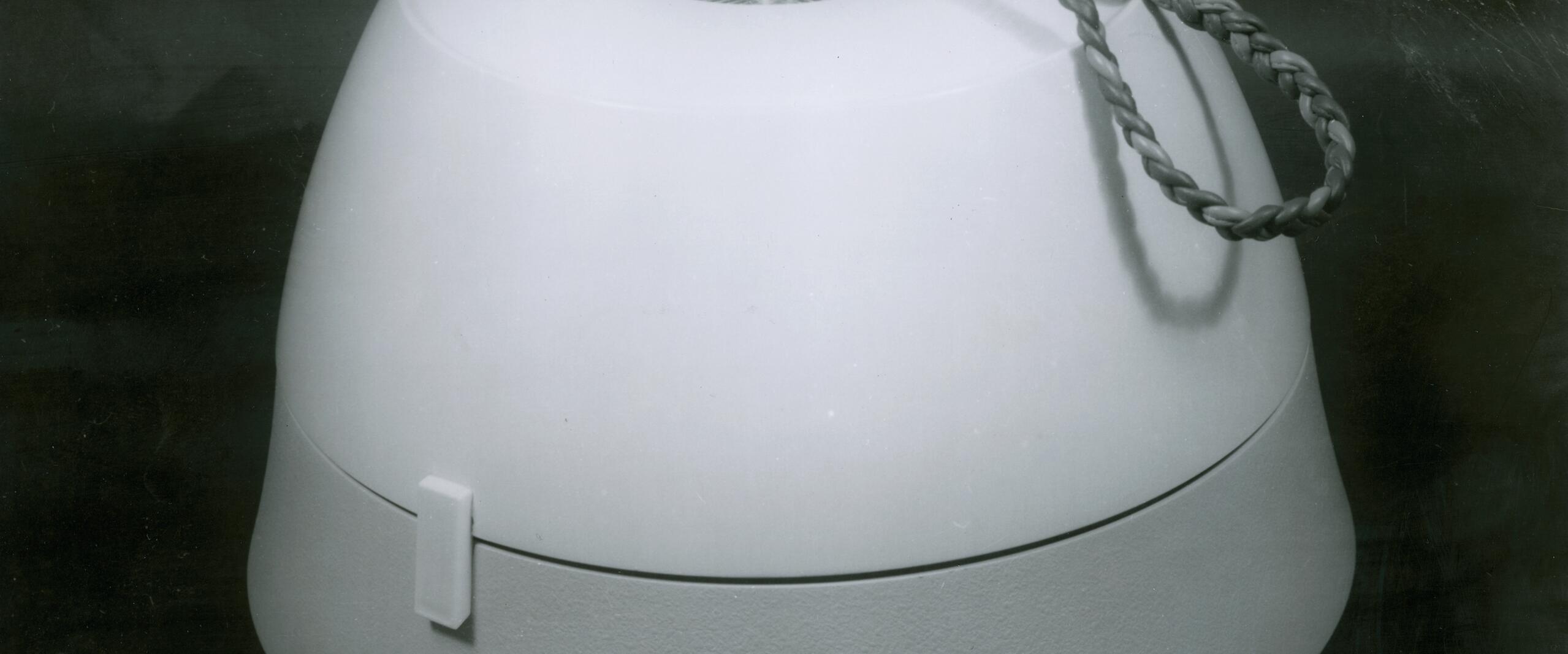 A round, smooth object with a rope handle on top, set against a dark background. The surface appears glossy with a small latch on the side.