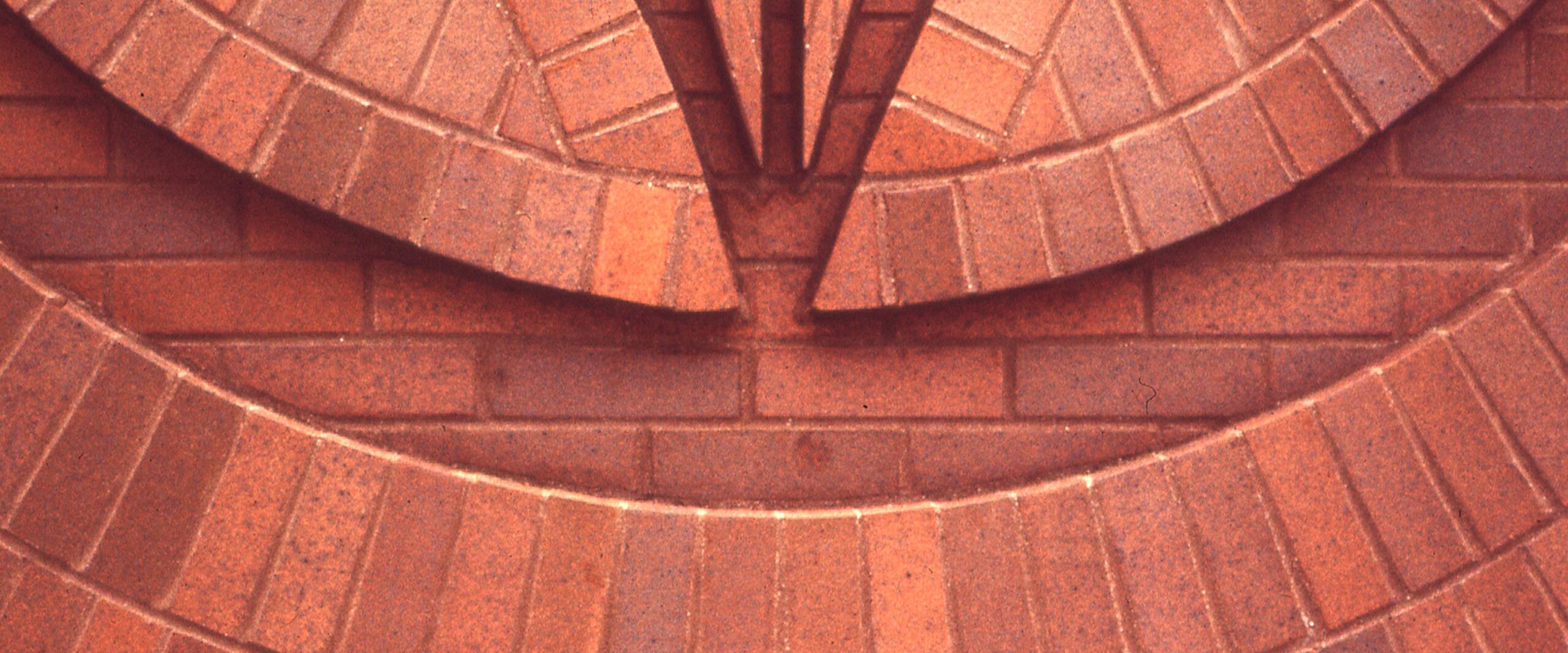 Abstract geometric pattern of interlocking red bricks forming concentric circular shapes and V-like angles.