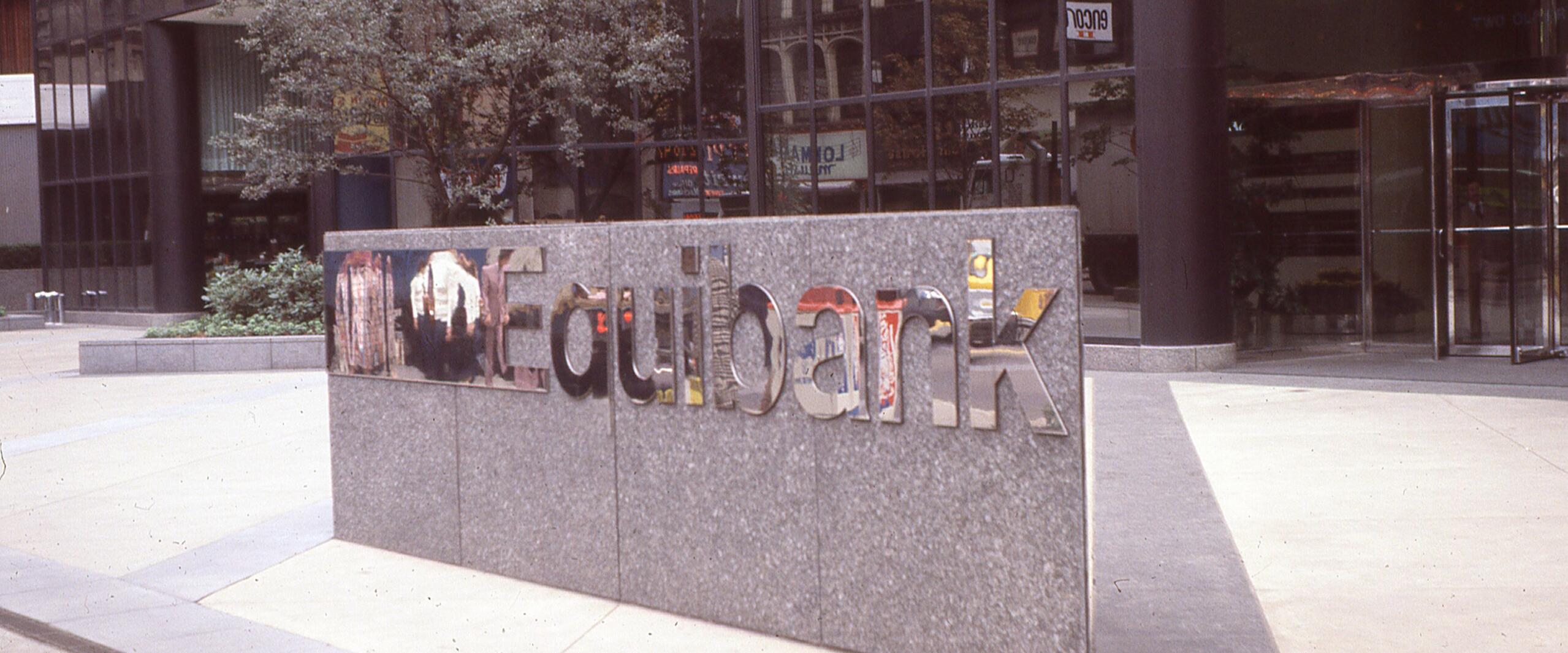 Granite wall with mirrored "Equibank" logo in an urban plaza, reflecting nearby buildings and trees.