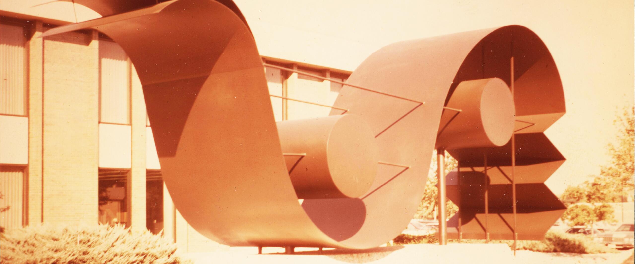 Large abstract metal sculpture with curved and circular elements, set in front of a brick building with vertical windows. Bushes are visible at the base. The image has a warm vintage tone.
