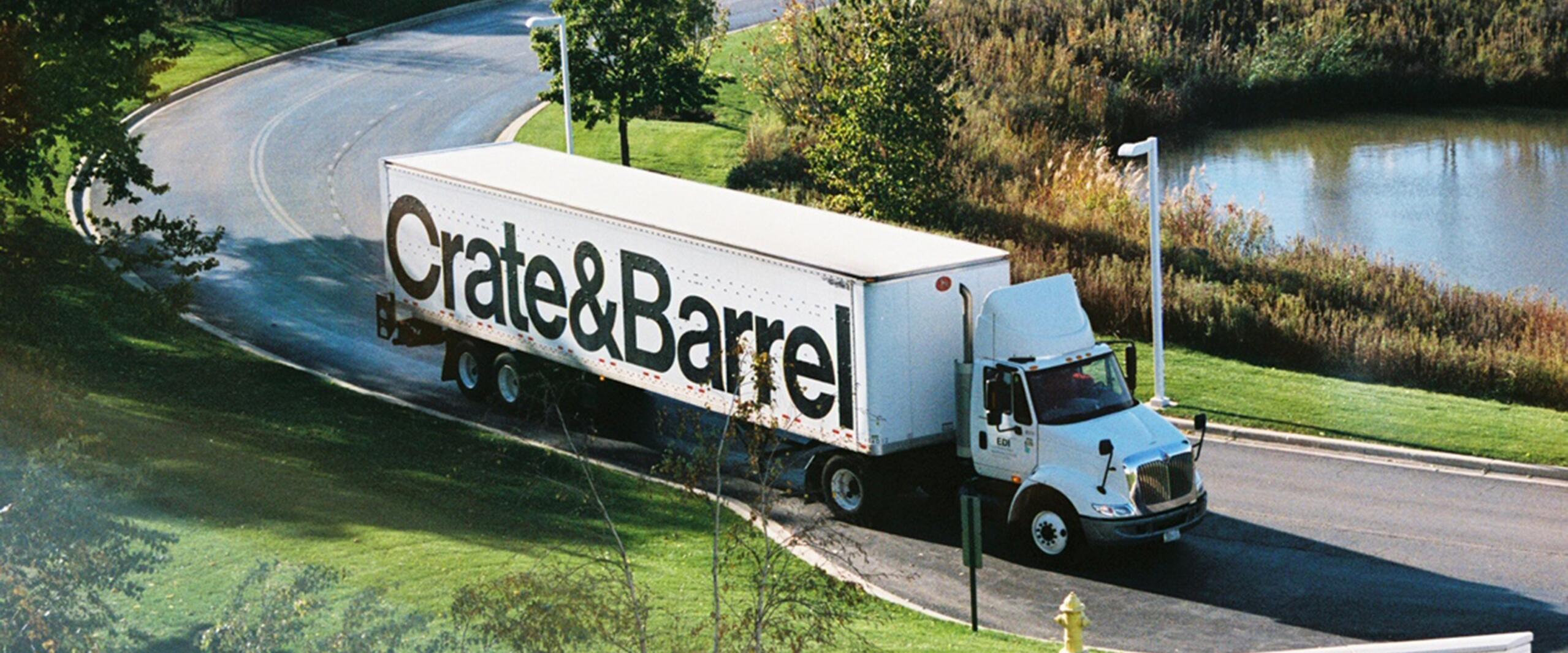 Truck with "Crate & Barrel" logo on the side driving on a curved road, surrounded by grass, trees, and a small pond, under clear blue skies.