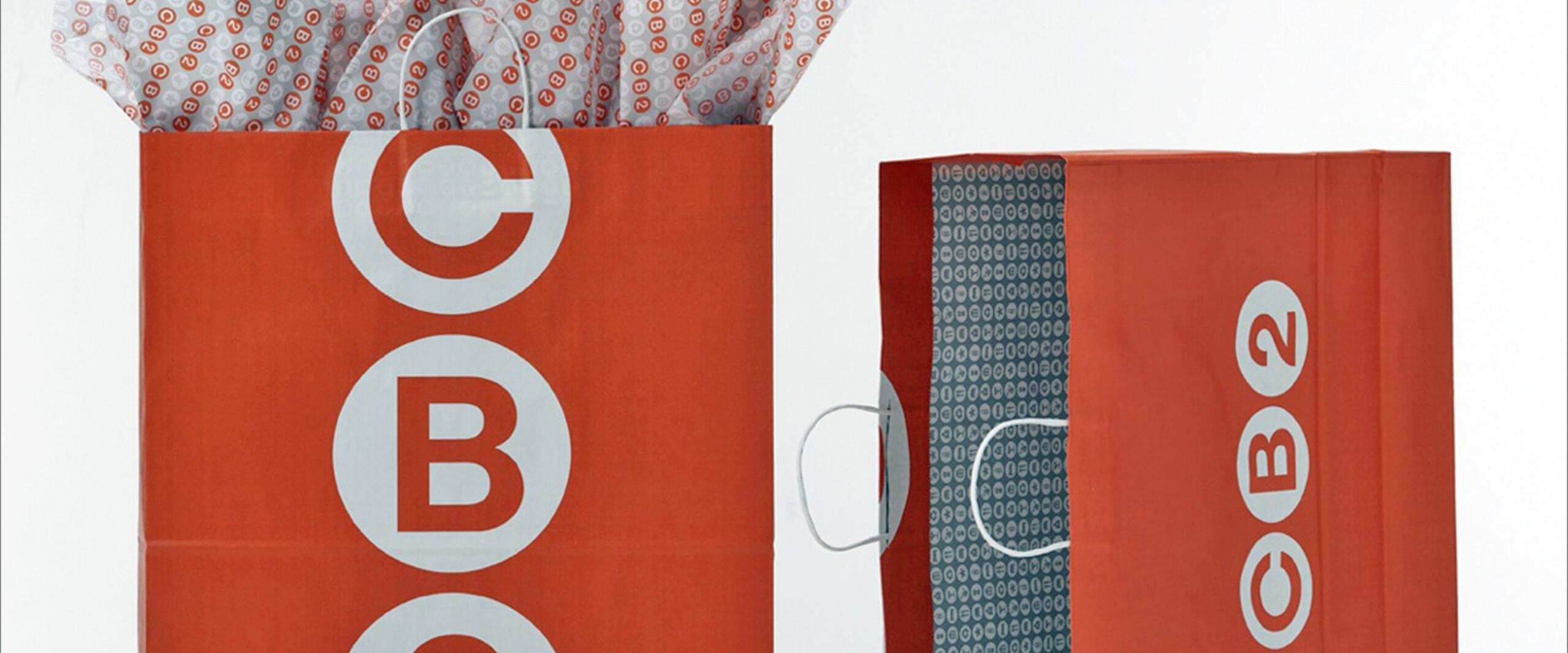 Two red shopping bags with white "CB2" logos. The bag on the left is filled with tissue paper, while the right bag is empty, displaying white handles and a patterned interior.