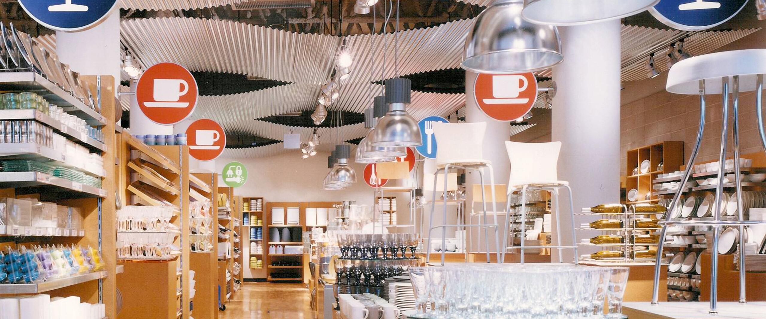 Store interior with wooden shelves and racks displaying dishes, glasses, and kitchenware. Hanging signs indicate sections with icons of wine glasses and teacups. Metal light fixtures hang from a wavy ceiling. Bright and organized layout.