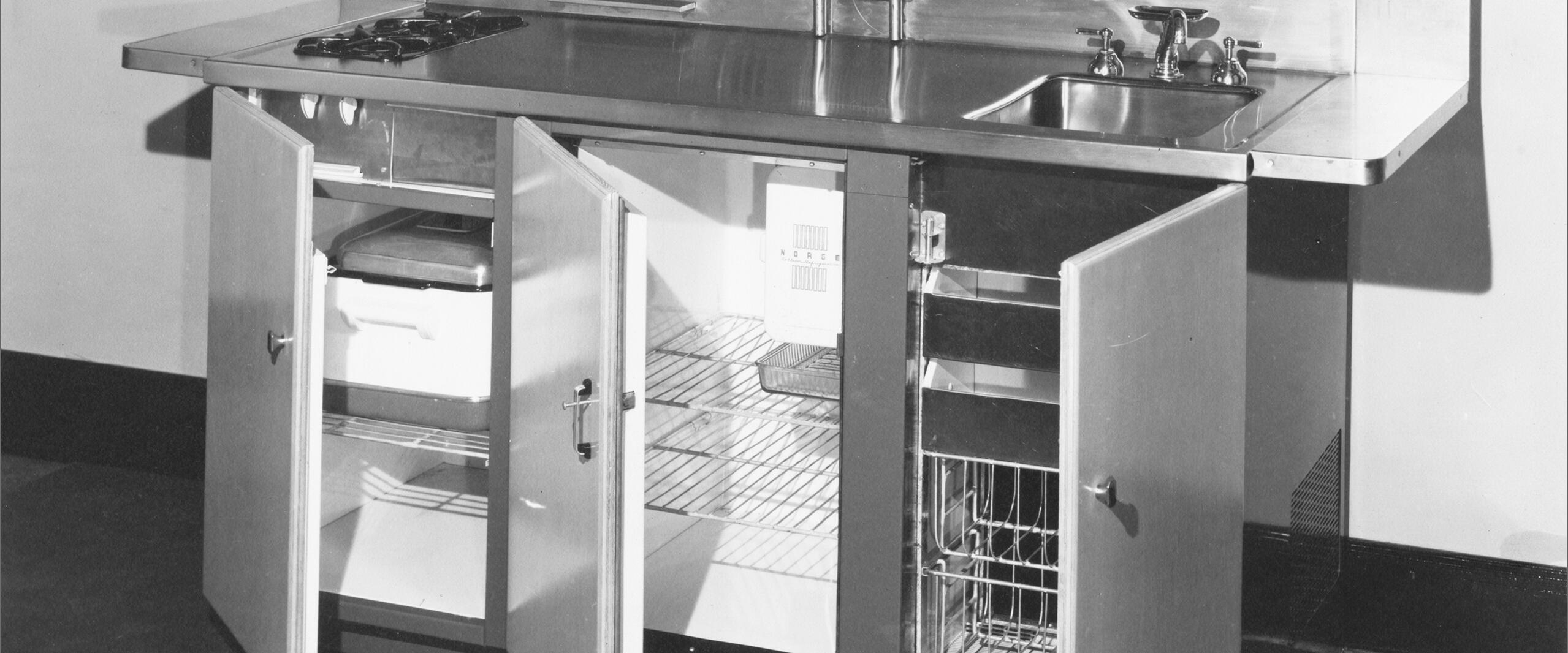Black and white photograph of a vintage kitchen unit with a stove, sink, and open wooden cabinet doors revealing shelves, a small fridge, and storage.