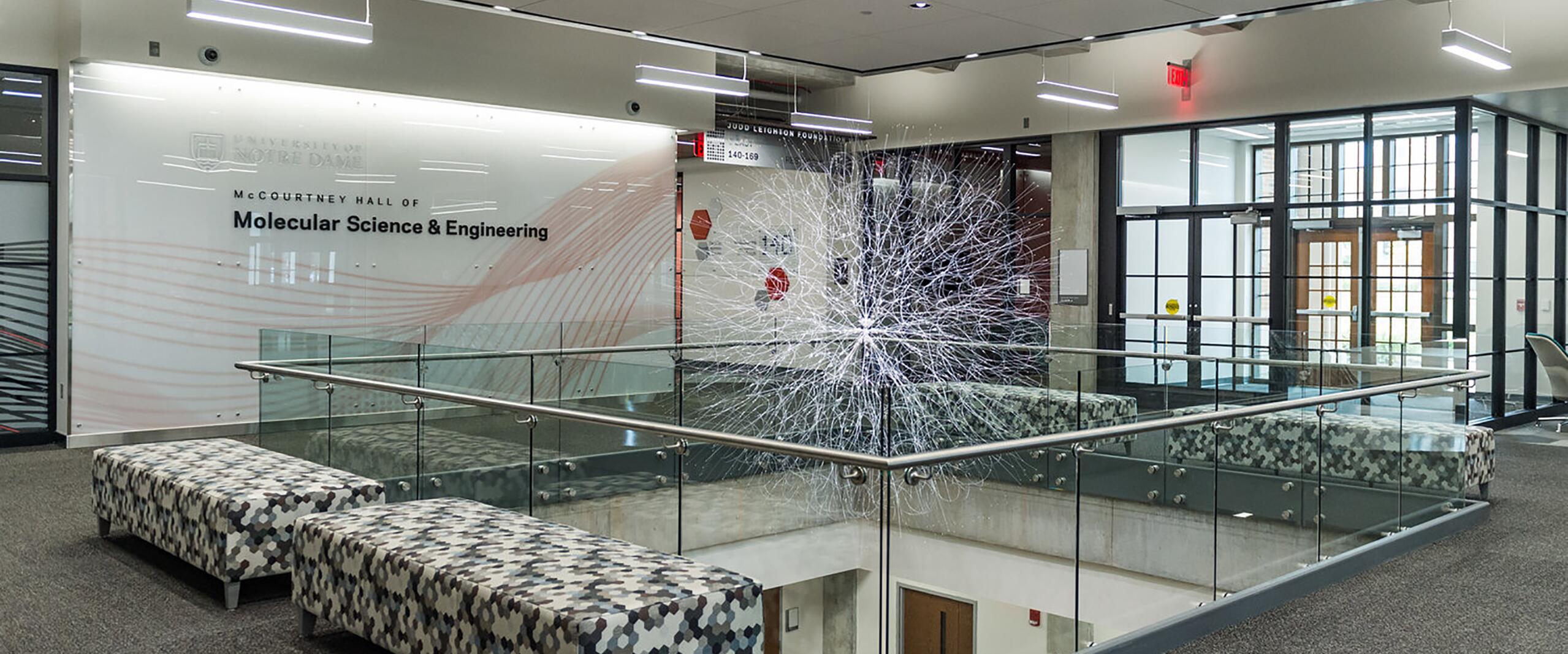 Modern hallway in McCourtney Hall of Molecular Science & Engineering, with glass railings, geometric-patterned benches, and a large abstract sculpture featuring a network of white lines.