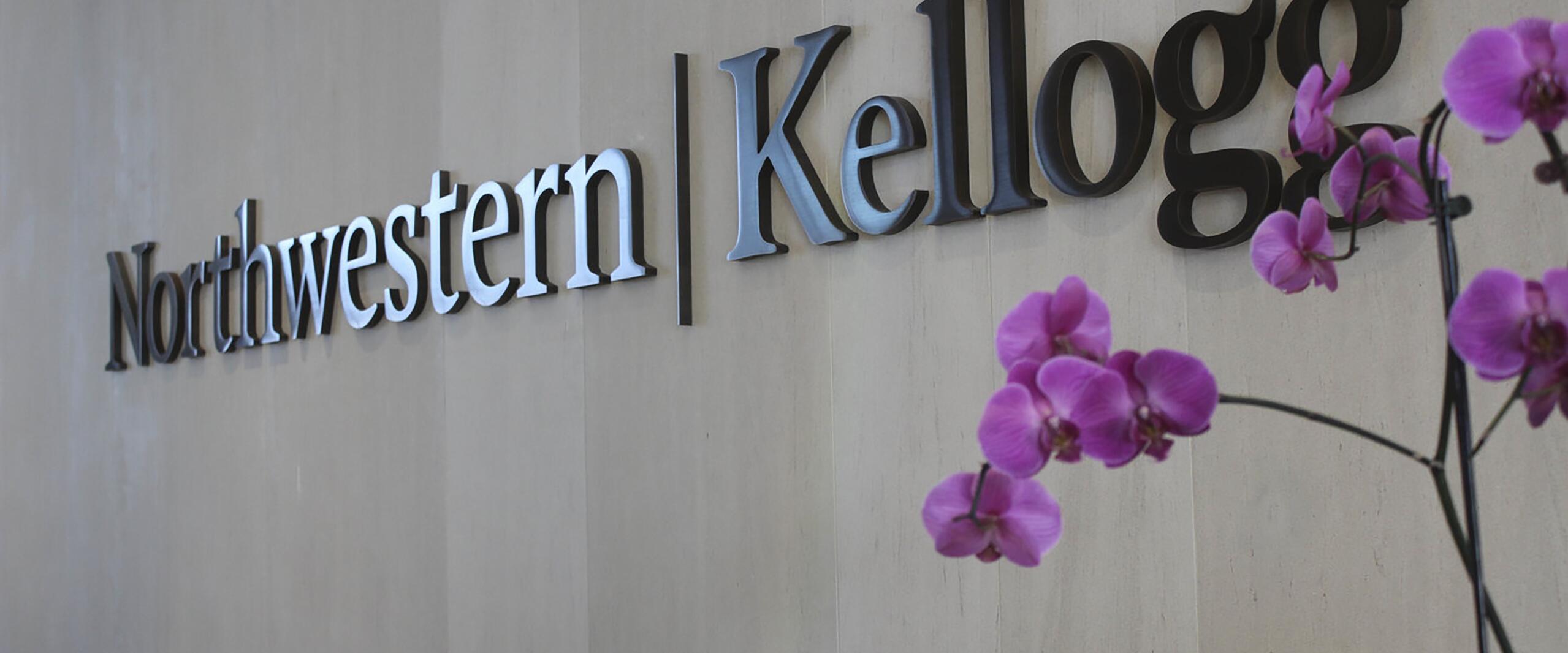 Northwestern Kellogg logo on a beige wall with purple orchids in the foreground.