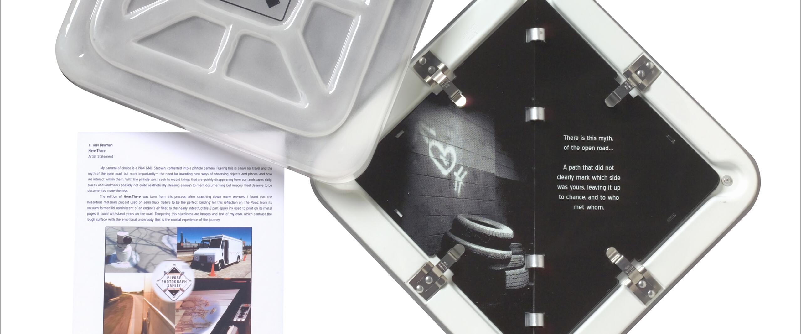 Open gray container with "Here There" logo and a booklet inside, featuring a photo of a tire stack against a wall and text about an open road. Nearby, a printed artist statement with smaller images, including a truck and landscapes.