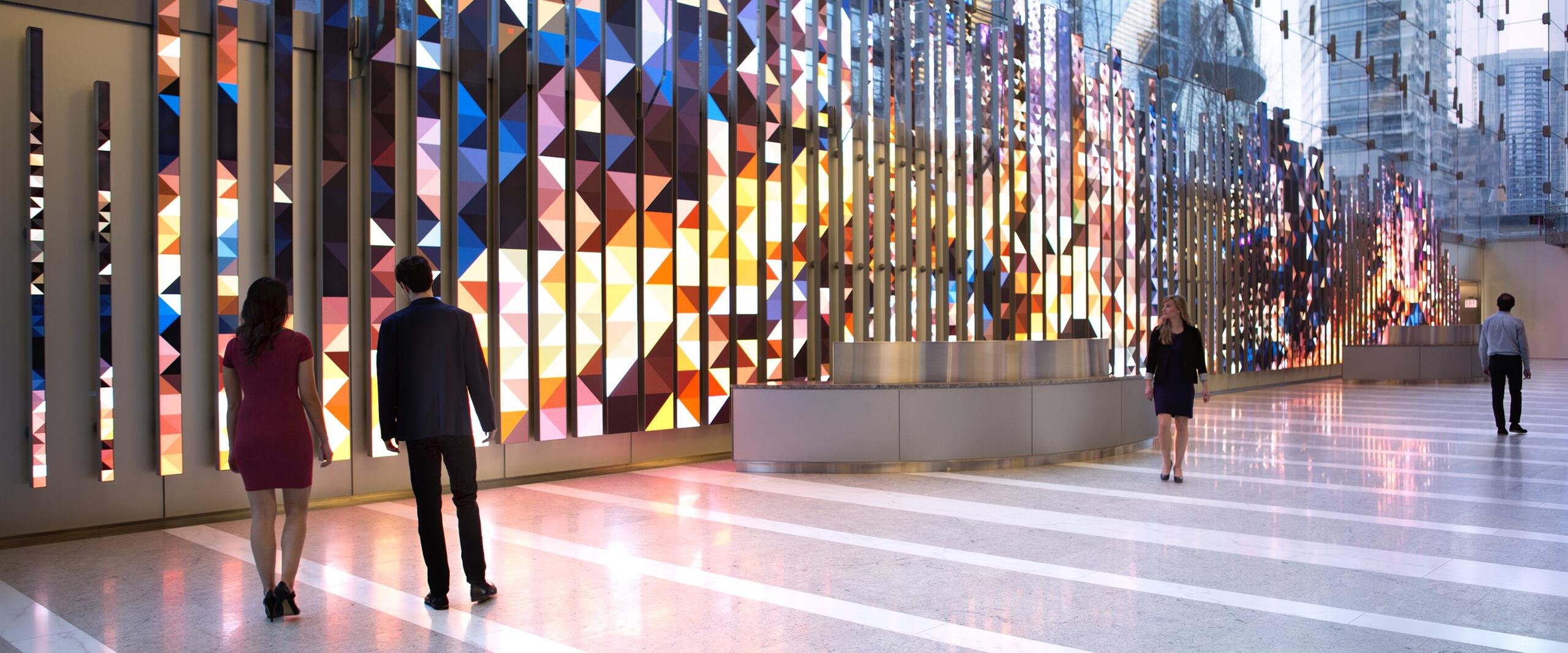 Colorful geometric wall art in a modern building lobby, reflecting vibrant patterns on the glossy floor. People walk along the illuminated corridor, with urban skyscrapers visible through glass windows.
