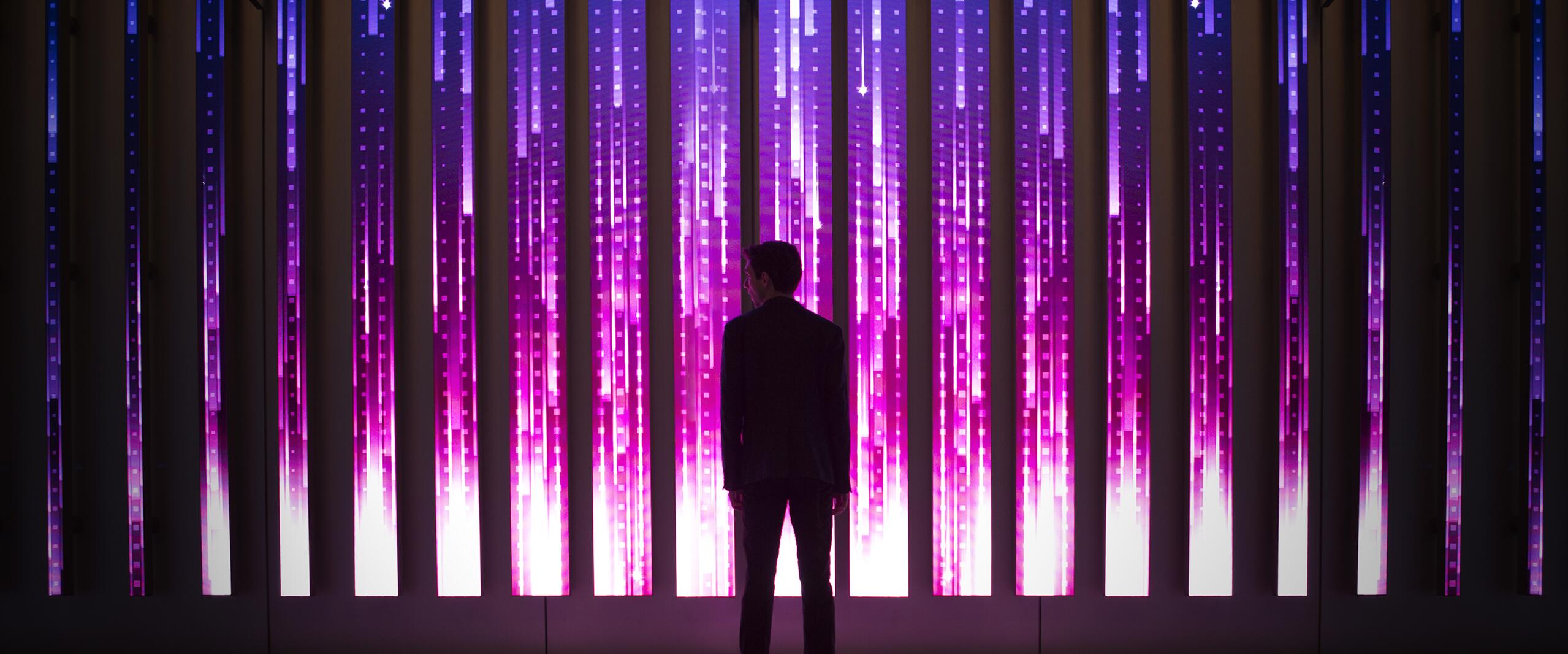 Silhouette of a person facing an illuminated wall with vertical blue and pink light strips, creating a futuristic, digital atmosphere.