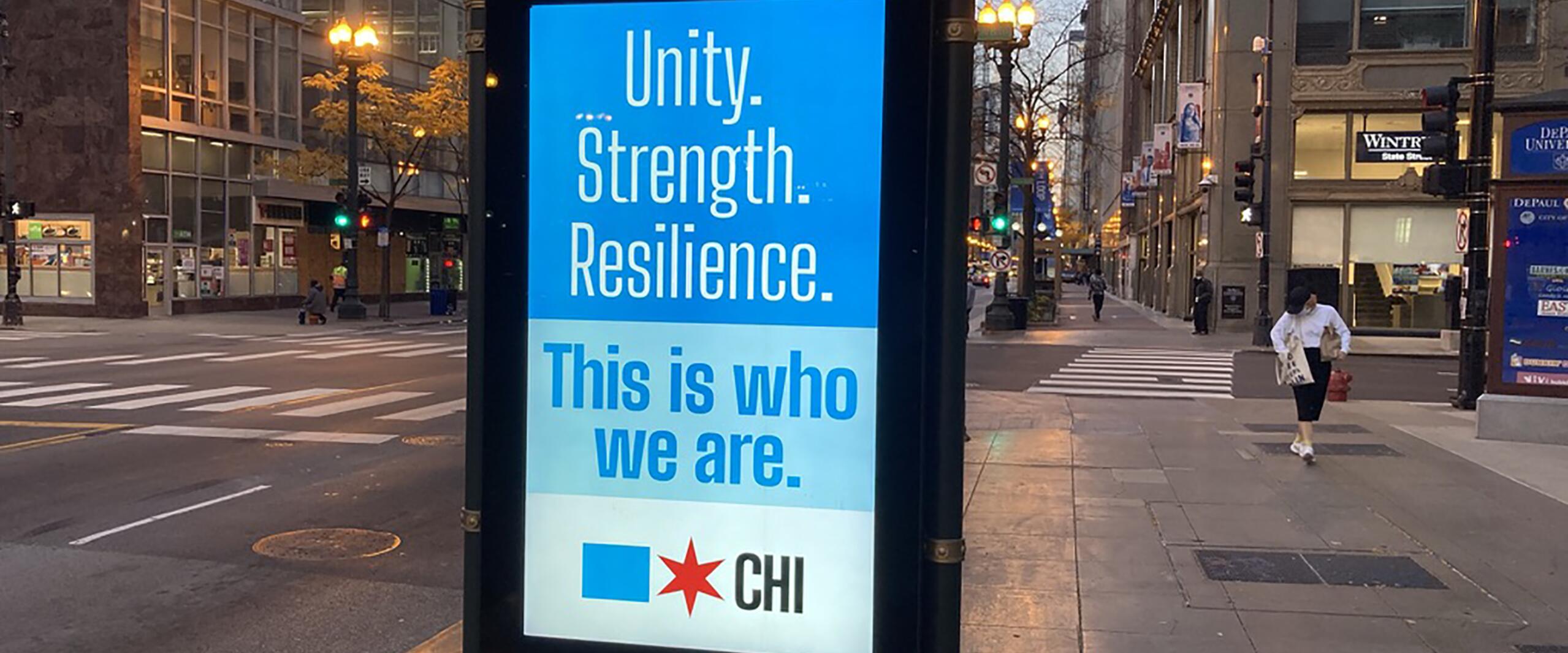 Street scene with a bus stop ad displaying a blue and white poster with the words "Unity. Strength. Resilience. This is who we are." The backdrop shows a city intersection and buildings illuminated by streetlights.