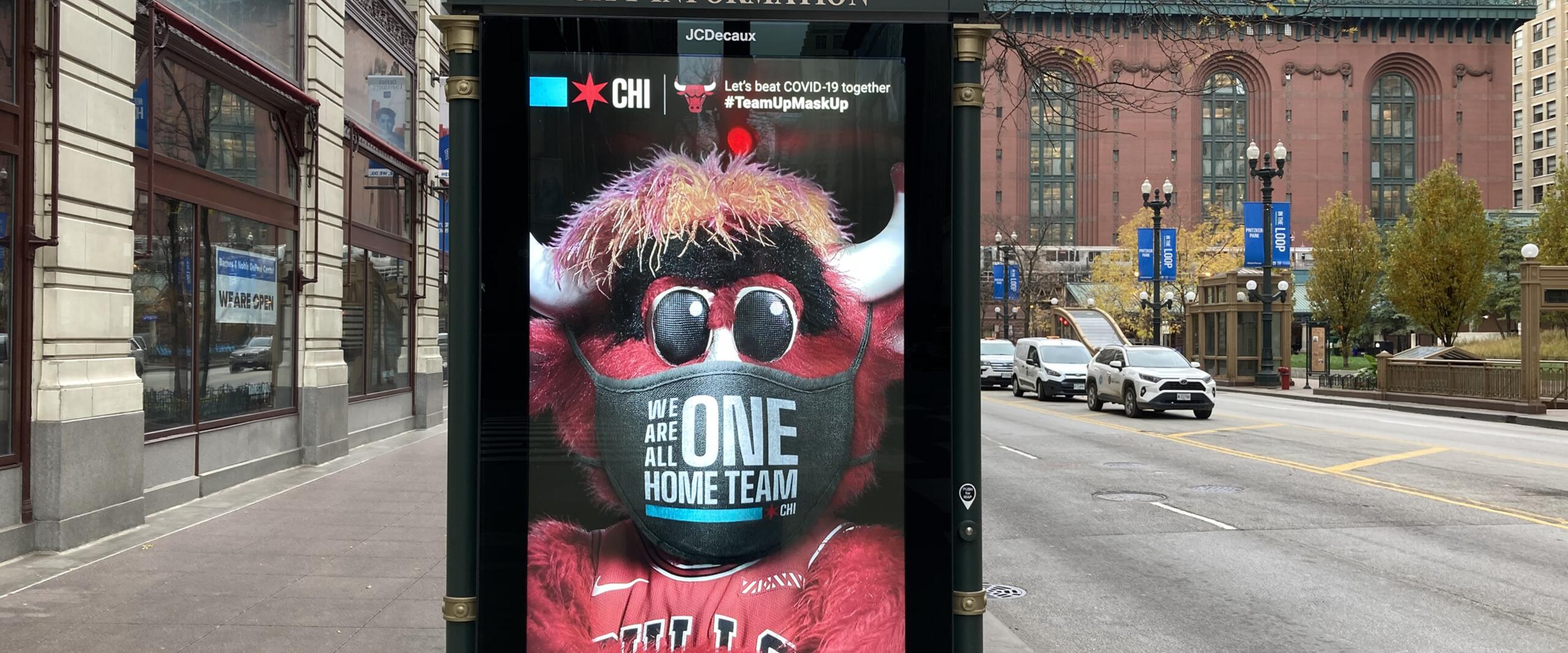 A street advertisement features a mascot wearing a mask with the phrase "We are all one home team," set against a city backdrop with historic buildings.