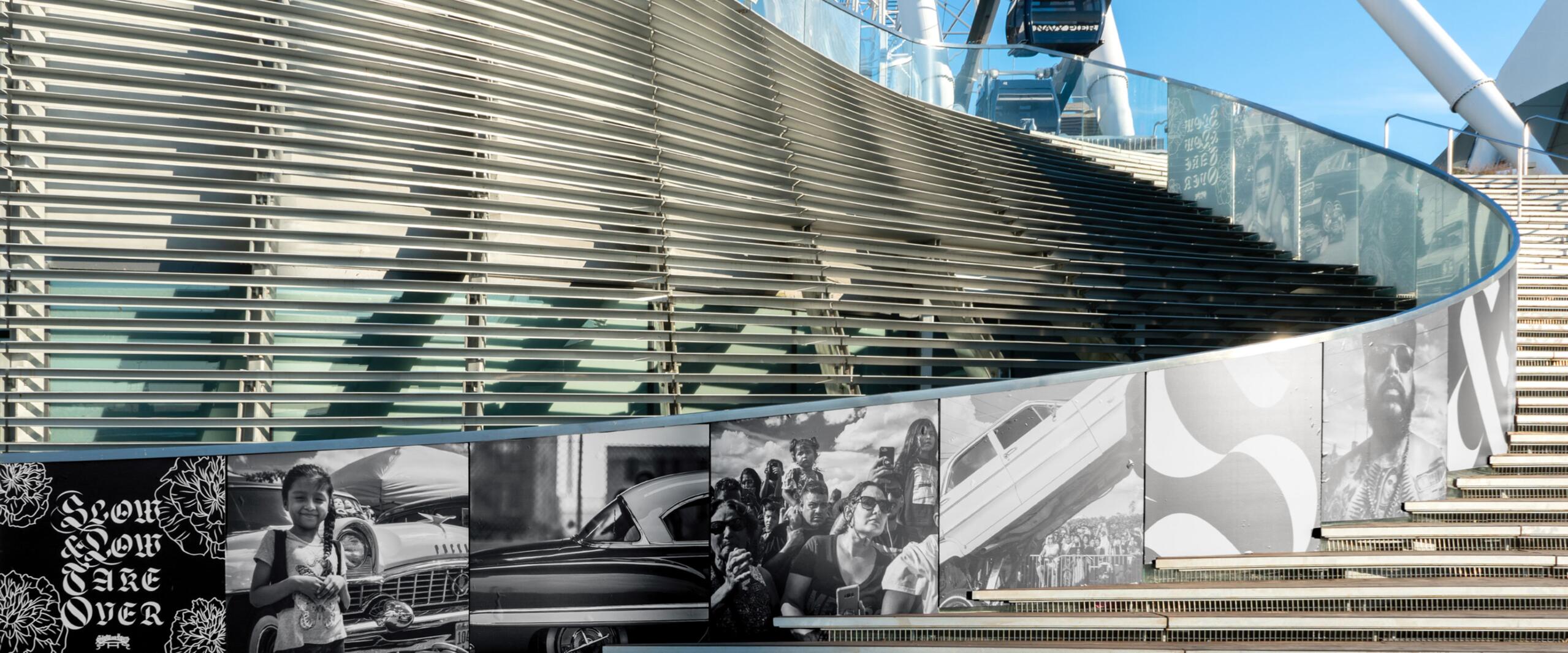 Curved staircase with metal railings, adorned with black and white murals. Background shows the structure of a Ferris wheel under a clear blue sky.