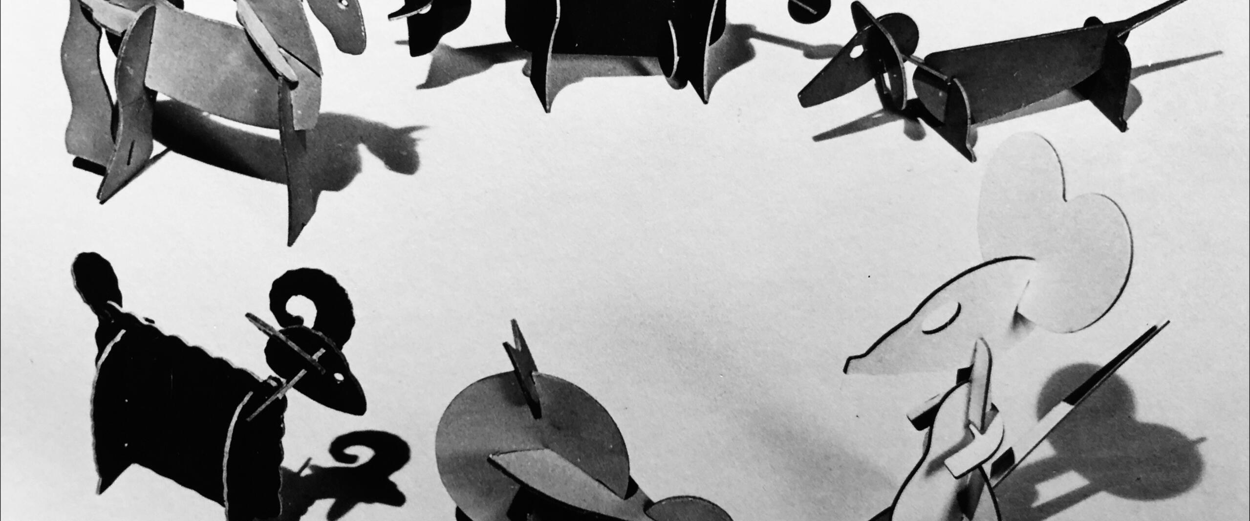 Paper animal models in various shapes including a unicorn, cow, dog, ram, duck, and mouse arranged in a circle on a flat surface, with shadows casting a playful pattern. Black and white photograph.