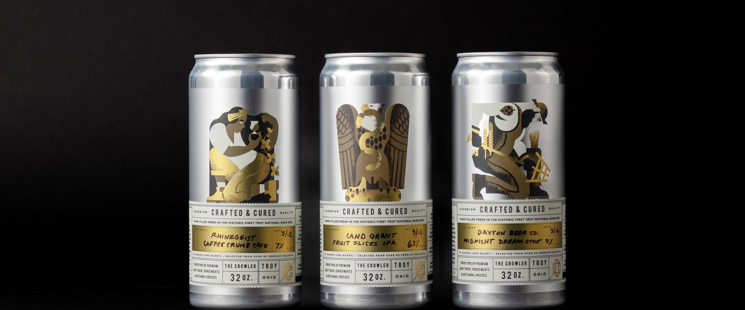 Three silver beverage cans with artistic gold labels featuring unique illustrations on a black background.