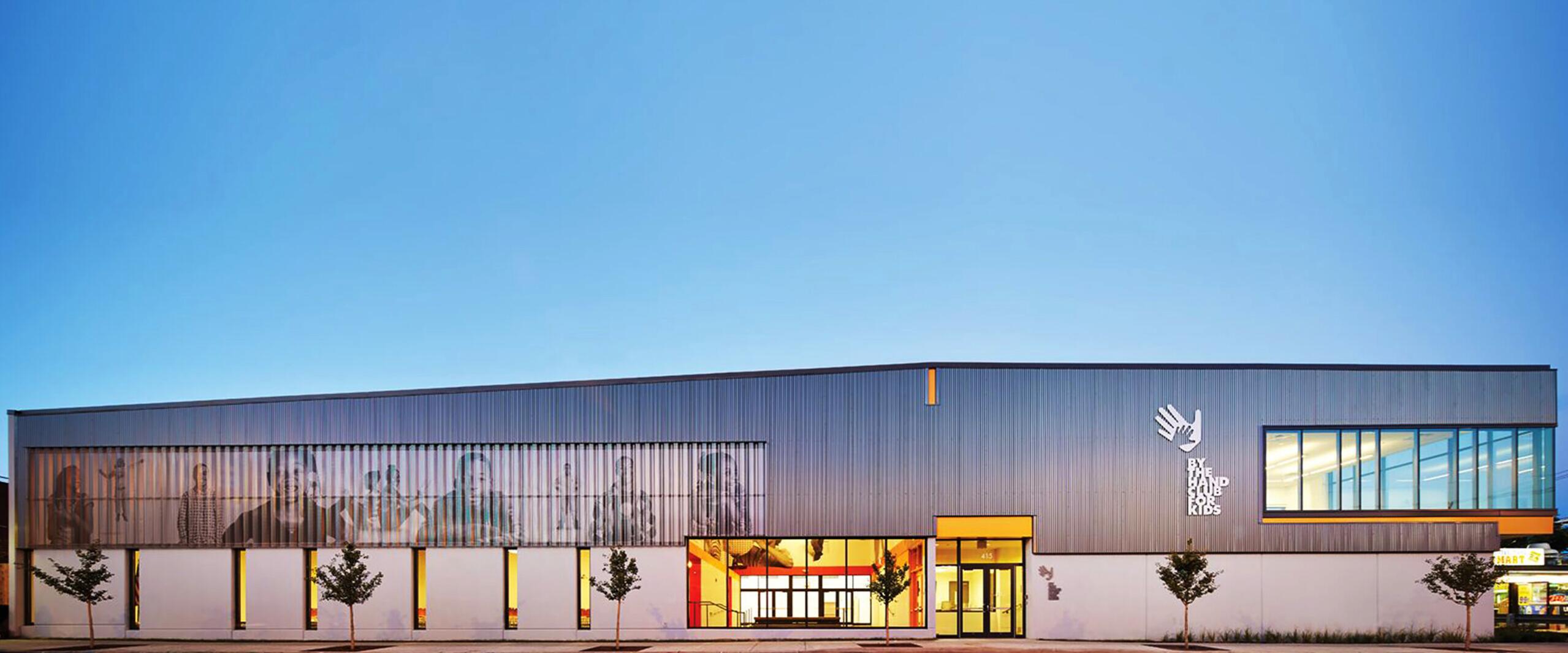 Modern building with a sleek, elongated facade, featuring large windows and a transparent section. The exterior is lit warmly from within, framed by a clear sky. Small trees line the sidewalk.