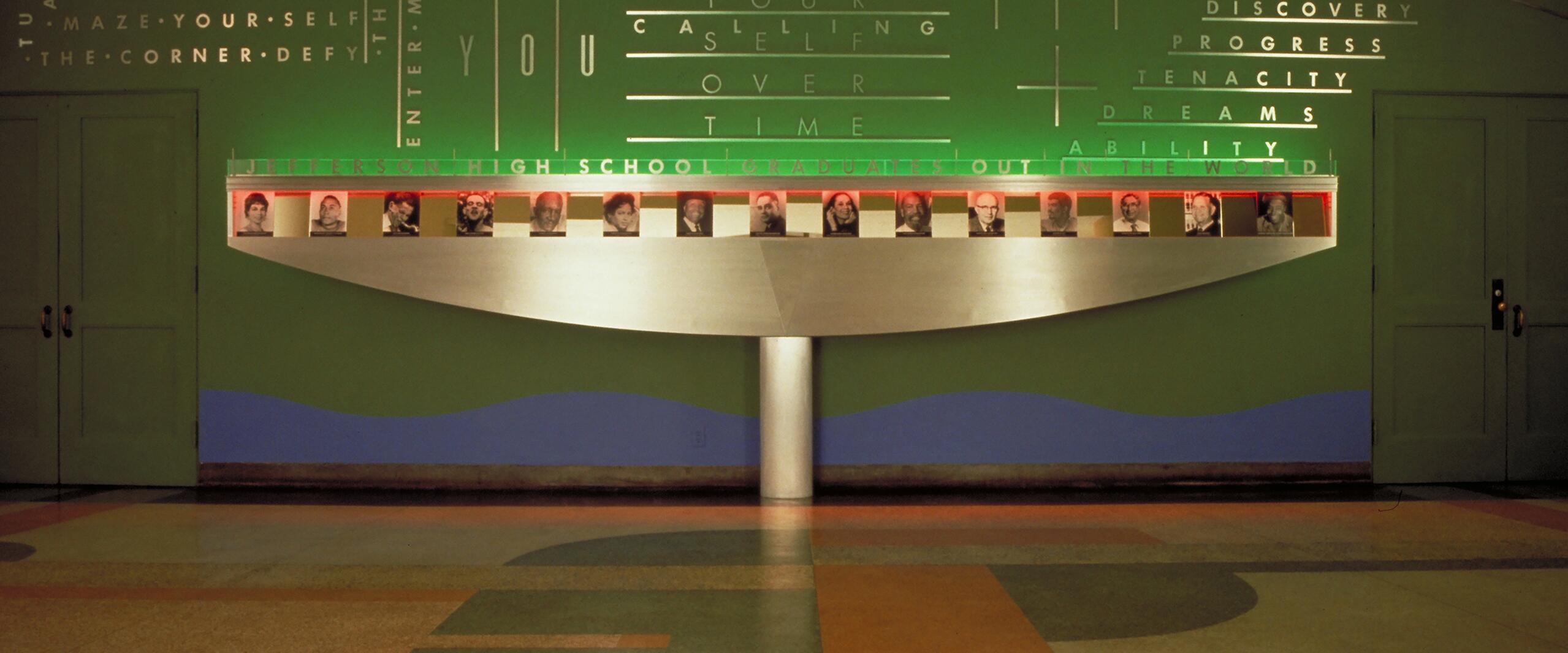 Colorful hallway featuring a green and blue wall with illuminated words and a display with framed photos of various people, flanked by two closed doors. The floor has a geometric pattern in orange, green, and tan.