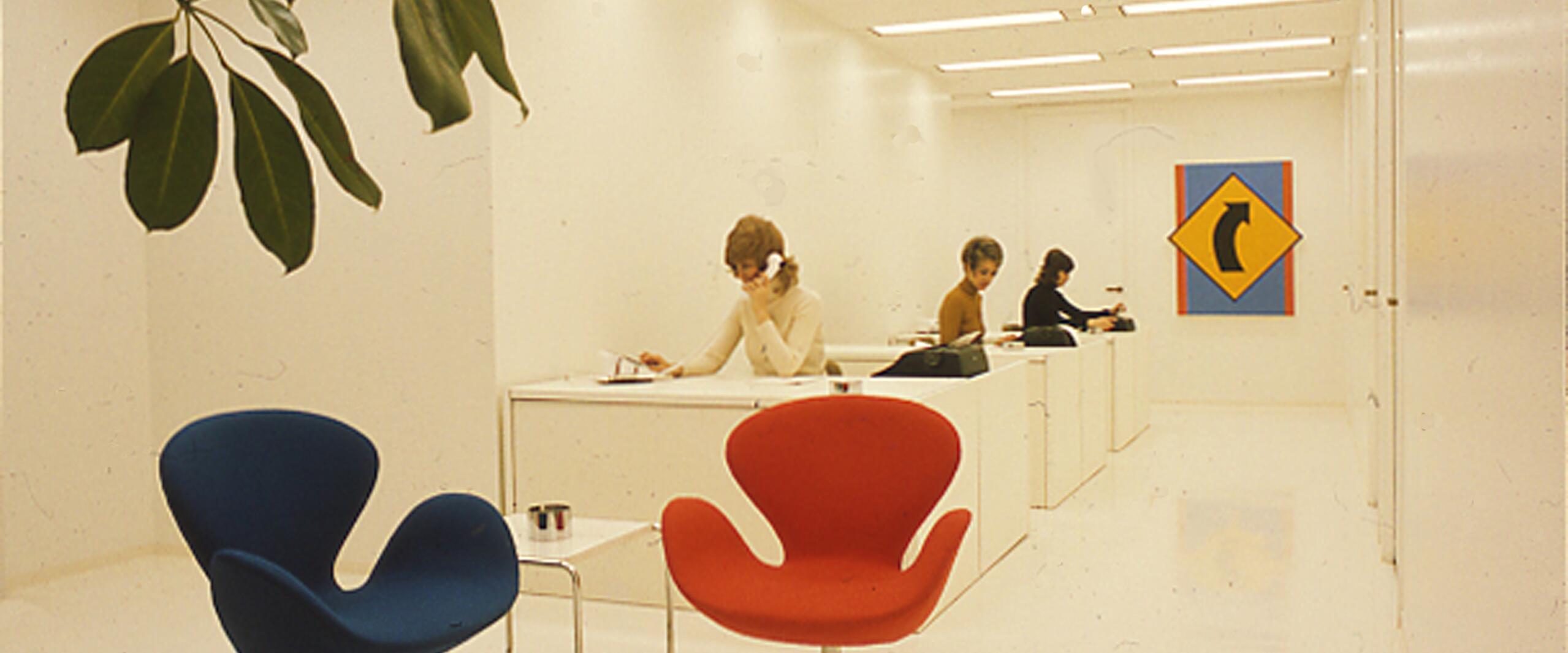 Minimalist office with white walls, a blue and a red mid-century modern chair, and three people working at desks in the background. A colorful abstract artwork hangs on the wall. A leafy plant is in the foreground.