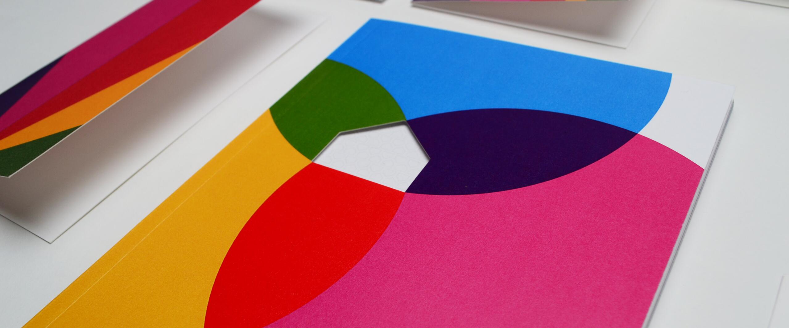 Colorful geometric patterns on white paper, featuring overlapping circles in blue, pink, red, yellow, green, and purple. Some sheets are folded and arranged on a flat surface.