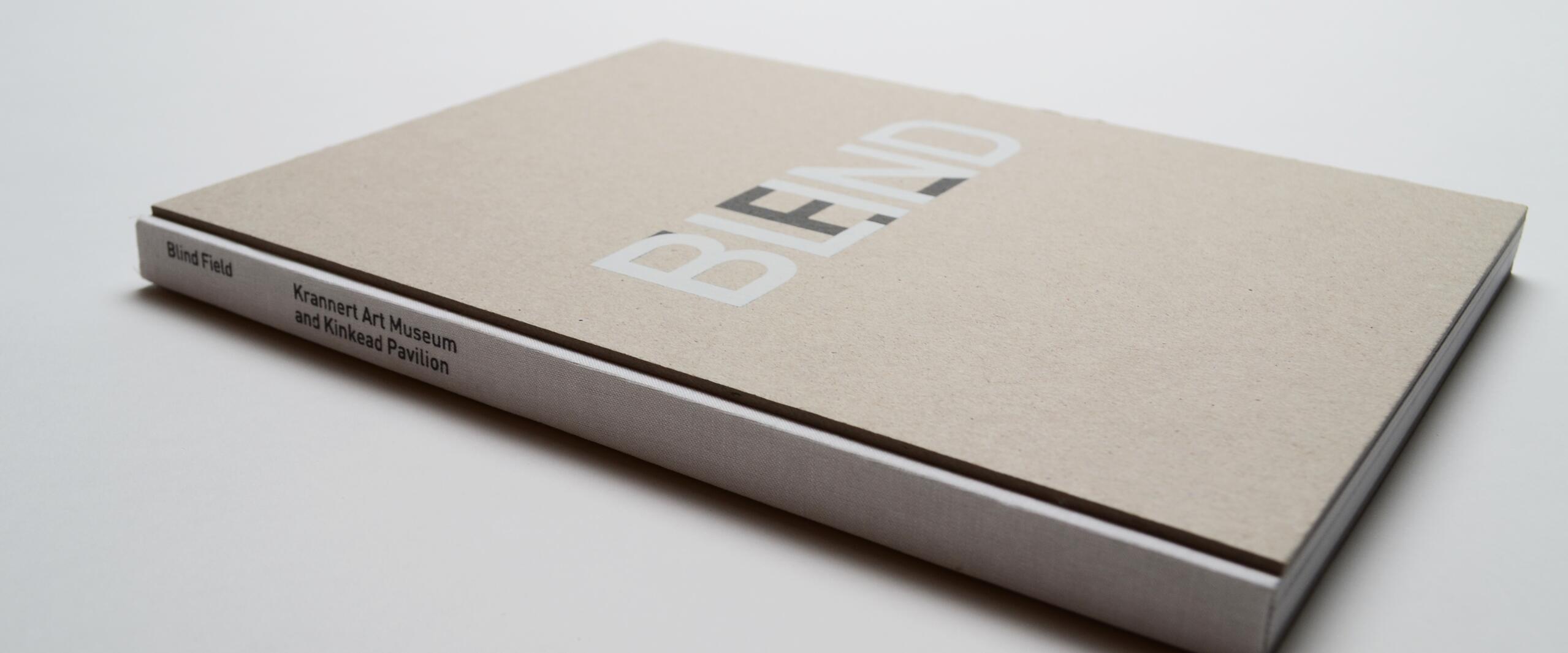 Tan hardcover book titled “BLEND” with white spine text “Blind Field, Krannert Art Museum and Kinkead Pavilion,” on a white surface.
