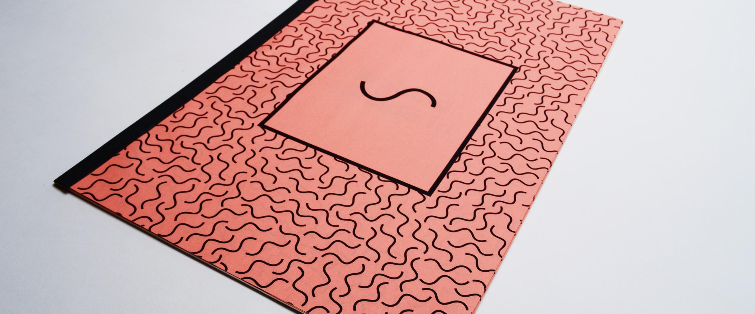 Orange notebook with wavy black lines, a black spine, and a large central rectangle featuring a single 'S' in the middle.