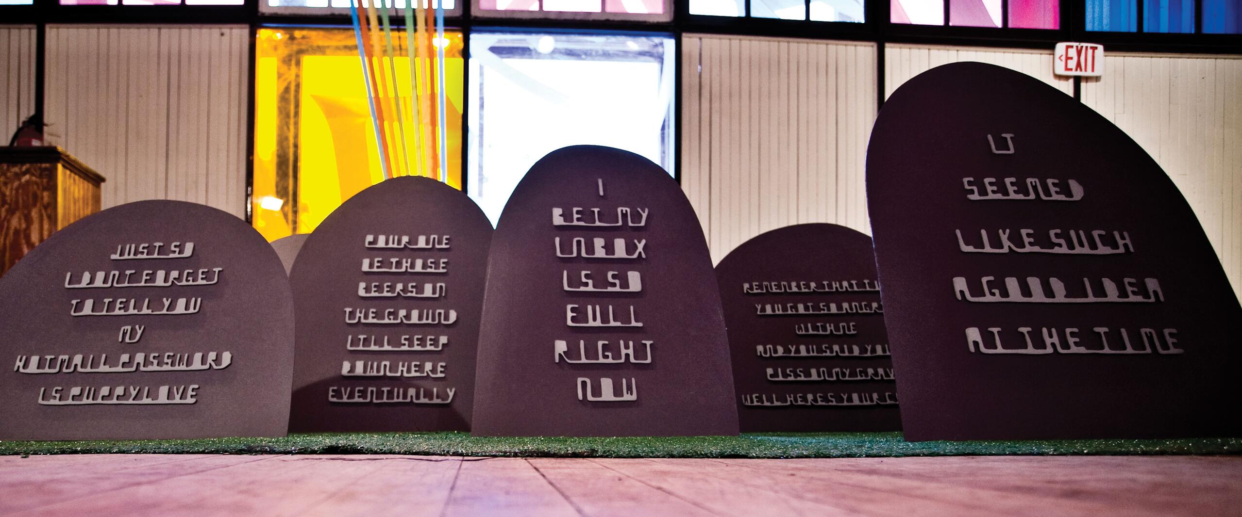 Art installation of tombstone-like structures with messages in cut-out letters, set in a colorful, illuminated indoor space with tall windows in the background.