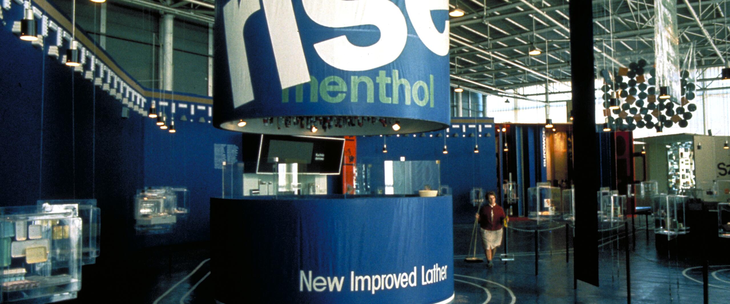 Exhibition booth with large blue cylindrical display featuring the word "rise" and "menthol," surrounded by modern exhibits and a person walking on a polished blue floor inside a spacious industrial hall with exposed metal beams.