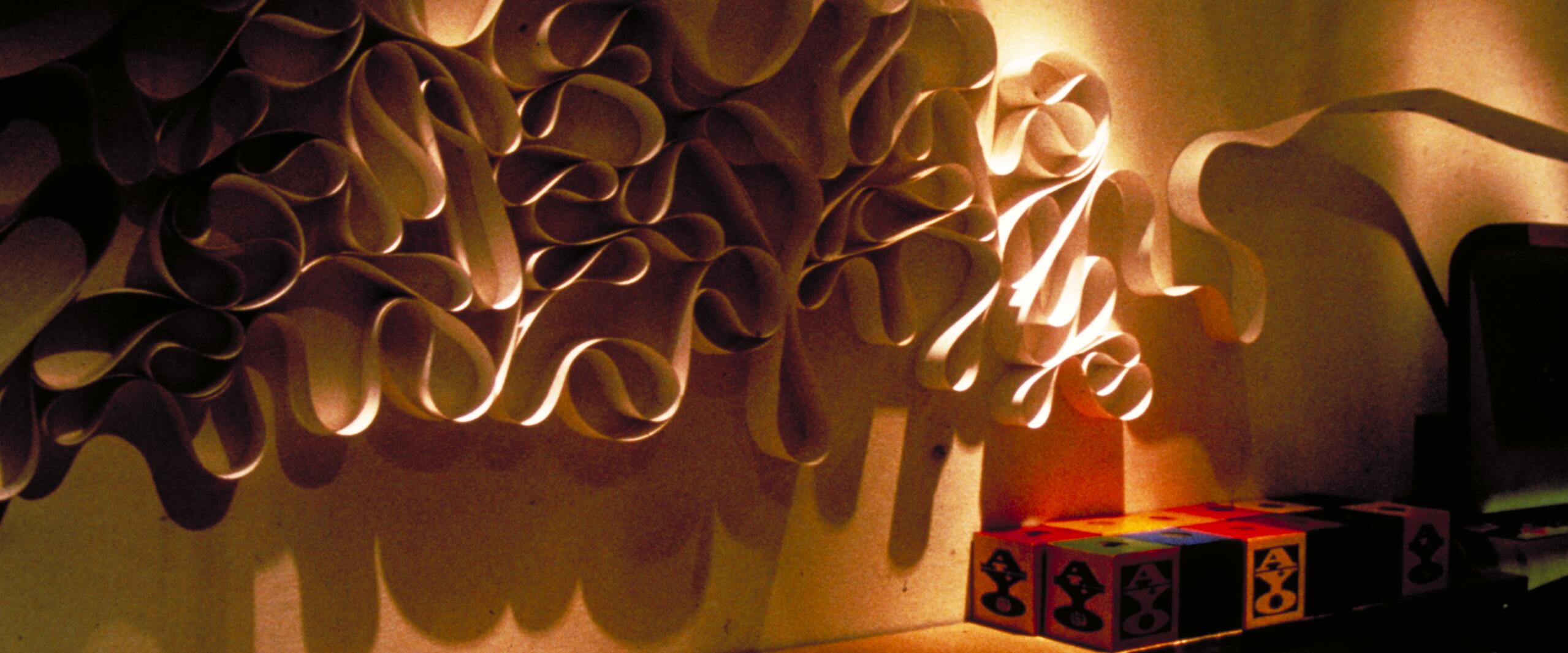 Abstract paper sculpture with intricate loops casting dramatic shadows on a wall, alongside colorful geometric cubes on a shelf, illuminated by warm lighting.