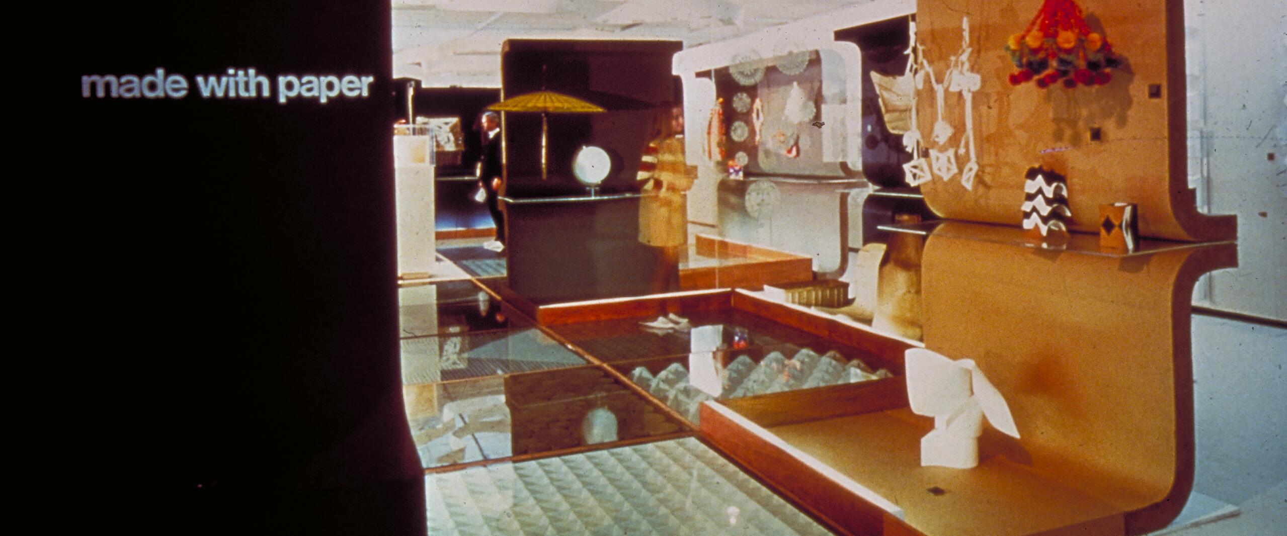 Exhibit showcasing various paper crafts and sculptures, displayed against a backdrop of curved, wooden panels. A soft, warm glow illuminates the scene, highlighting models and paper art.