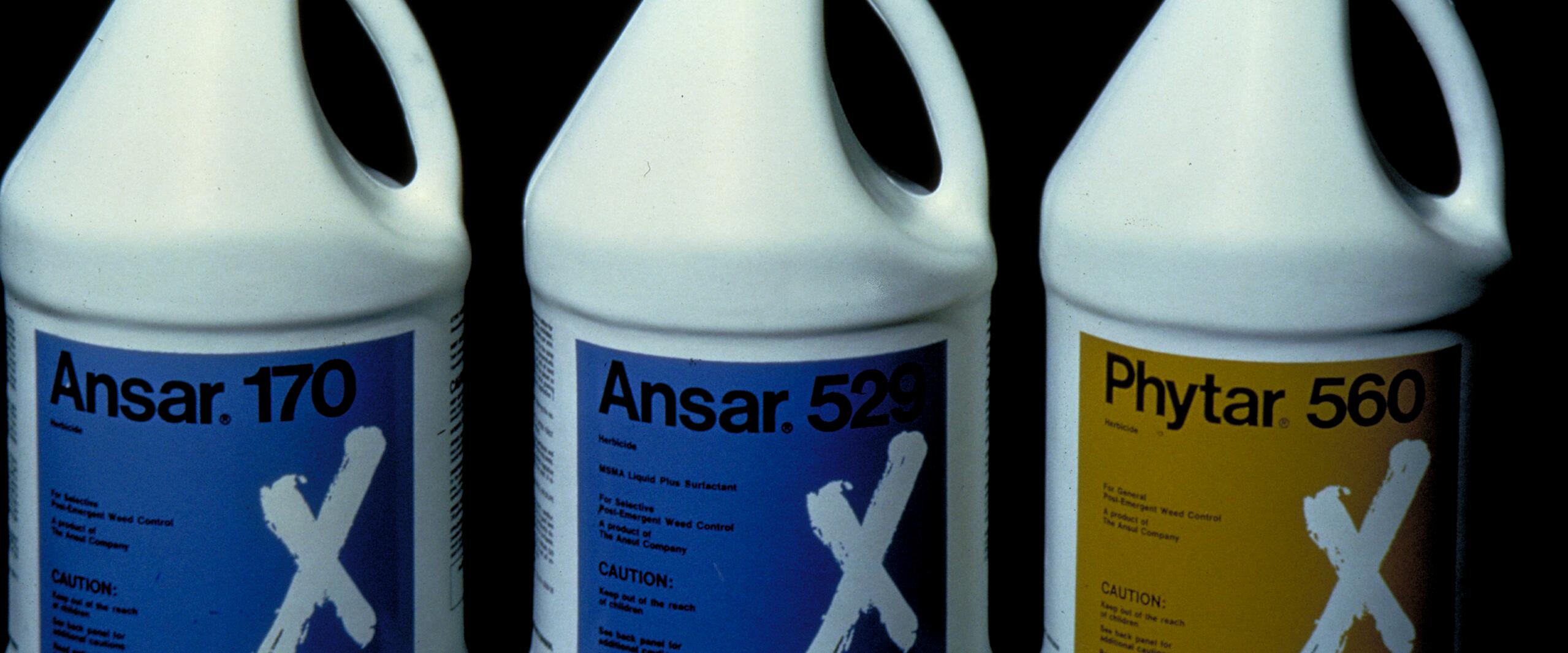 Three white gallon containers with handles, labeled "Ansar 170," "Ansar 529," and "Phytar 560." Labels have blue and yellow backgrounds with a large white "X." Containers are arranged side by side against a black background.
