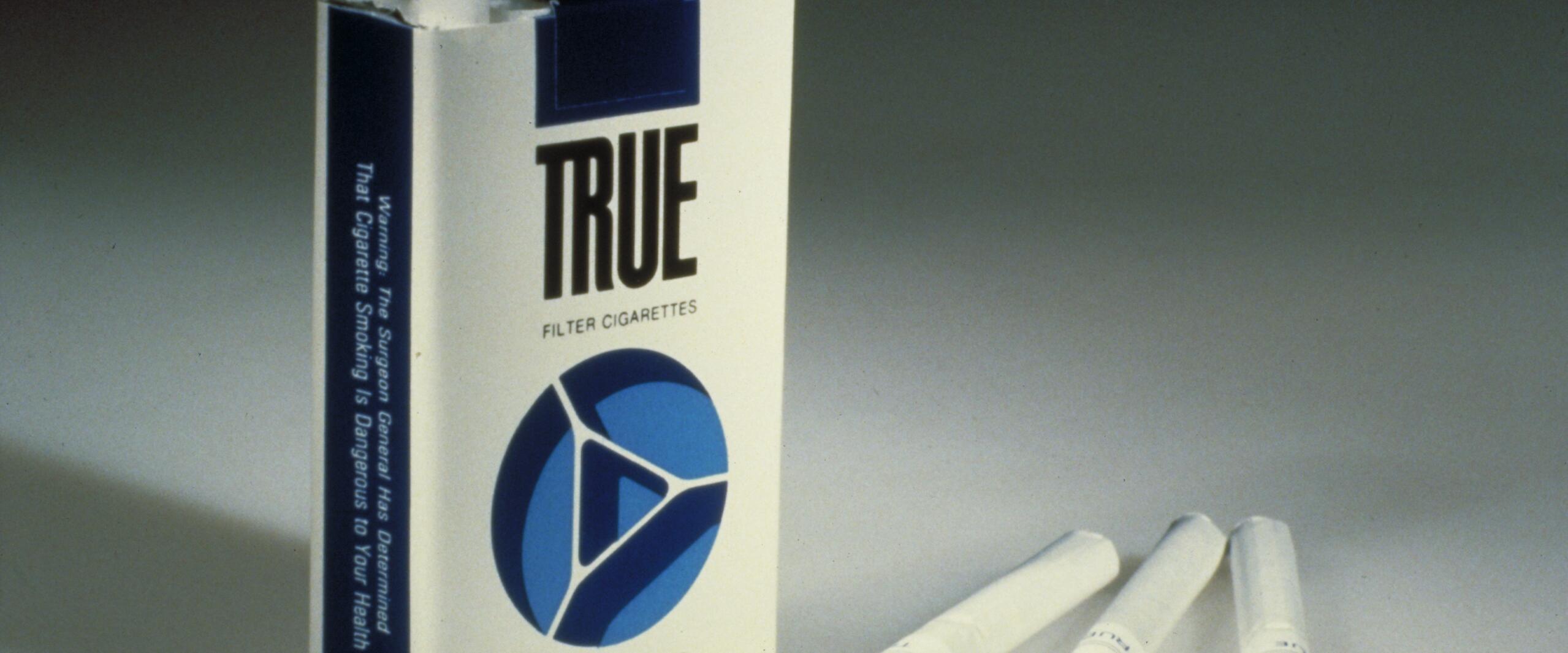 Cigarette pack labeled "TRUE Filter Cigarettes Ultra Low Tar" with two cigarettes lying beside it on a light surface.