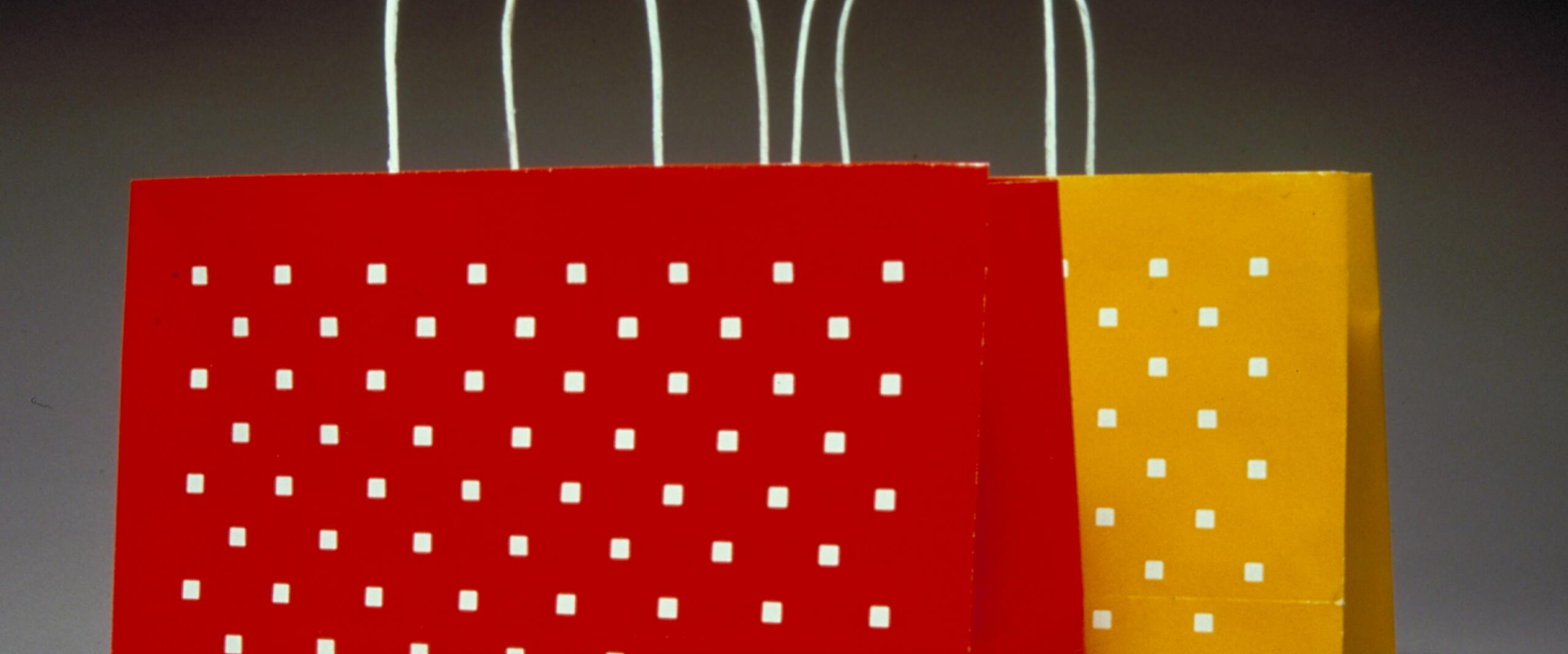Two paper shopping bags with white square patterns, one red and one yellow, stand side by side against a gradient dark background. Each bag has white handles.