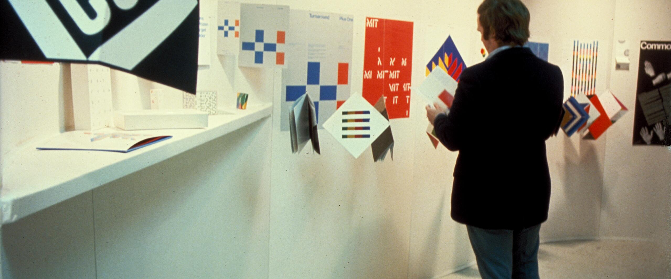Man viewing a display of colorful graphic design posters on a curved white wall, featuring geometric patterns and bold text.