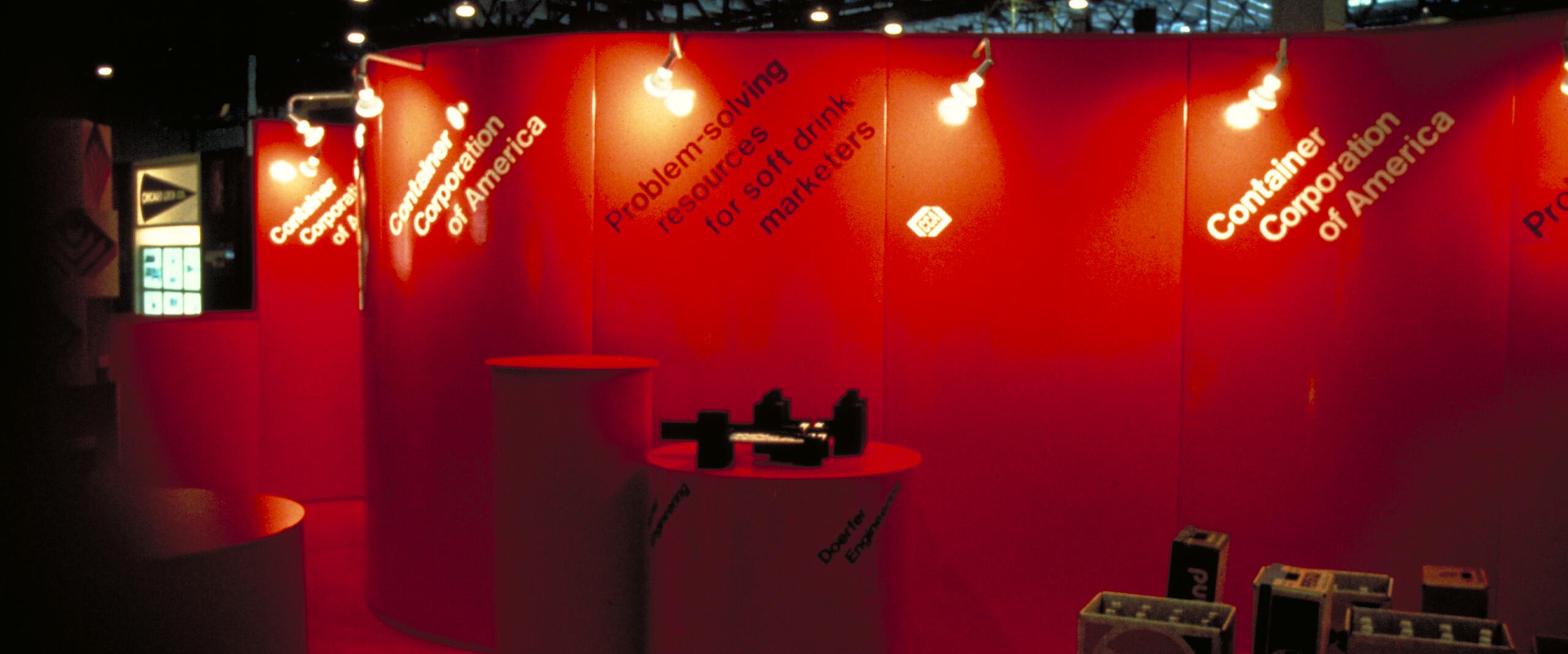 Exhibition booth with red panels and cylindrical display tables. Large white text highlights the Container Corporation of America. Bright lighting and product boxes, including Pepsi-branded items, are visible beneath the spotlights.